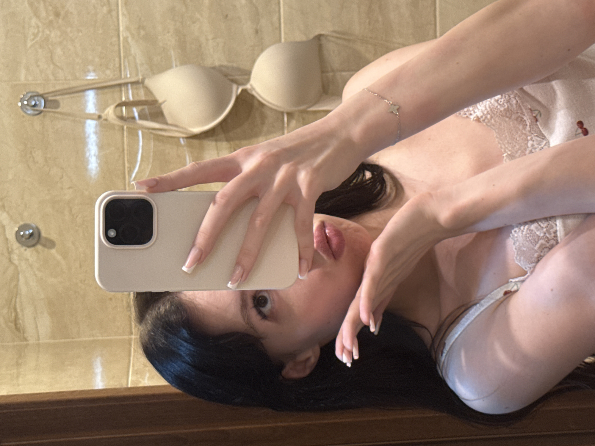 A young woman with long, dark hair takes a selfie in a bathroom. She is wearing a light-colored, lace-trimmed camisole. Her left hand holds a smartphone, obscuring part of her face, while her right hand is raised near her mouth in a playful gesture. A beige bra is hanging on hooks on the tiled wall behind her. The overall lighting is warm and diffused, suggesting indoor ambient light. There is no indication of the city of Khimki, Russia.