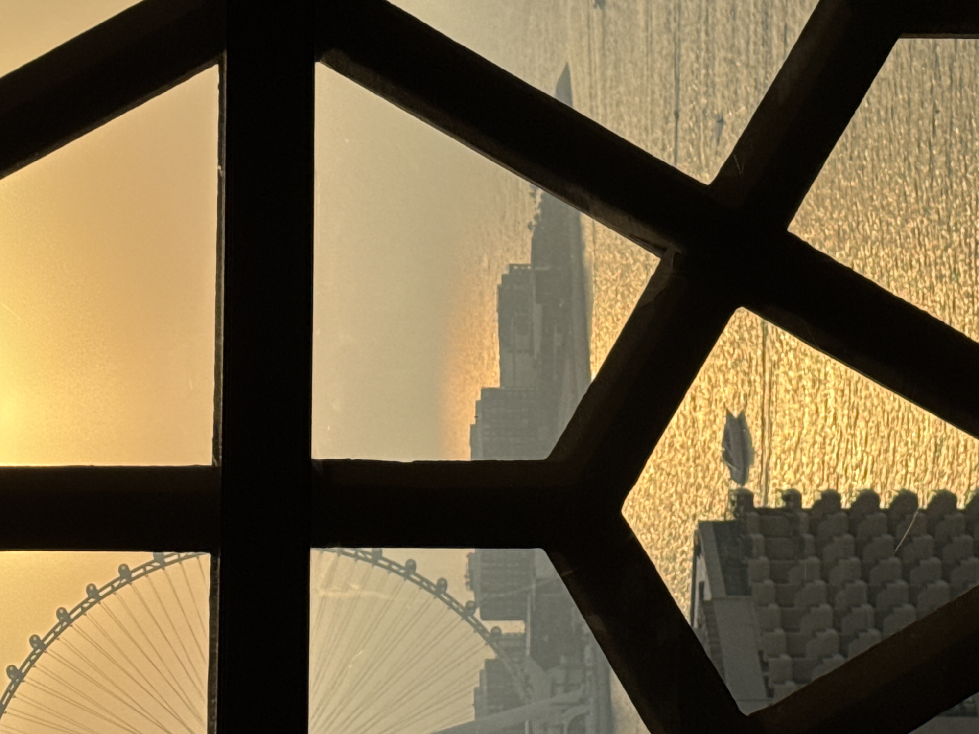 The image captures a view through a geometric frame, likely a window, overlooking a body of water in Dubai, United Arab Emirates, during sunset. The warm, golden light of the setting sun reflects off the water, creating a shimmering effect. In the distance, a large Ferris wheel, possibly the Ain Dubai, is partially visible on the left side, its spokes fanning out against the bright sky. A line of buildings, likely hotels or residential structures, are situated along the waterfront. A small boat is seen navigating the water in the middle ground. On the right side, the water stretches towards the horizon, with hints of other small boats. The foreground features a dark, angular frame that segments the view into different sections, adding a sense of depth and perspective. The overall scene evokes a serene and picturesque urban coastal landscape at dusk.