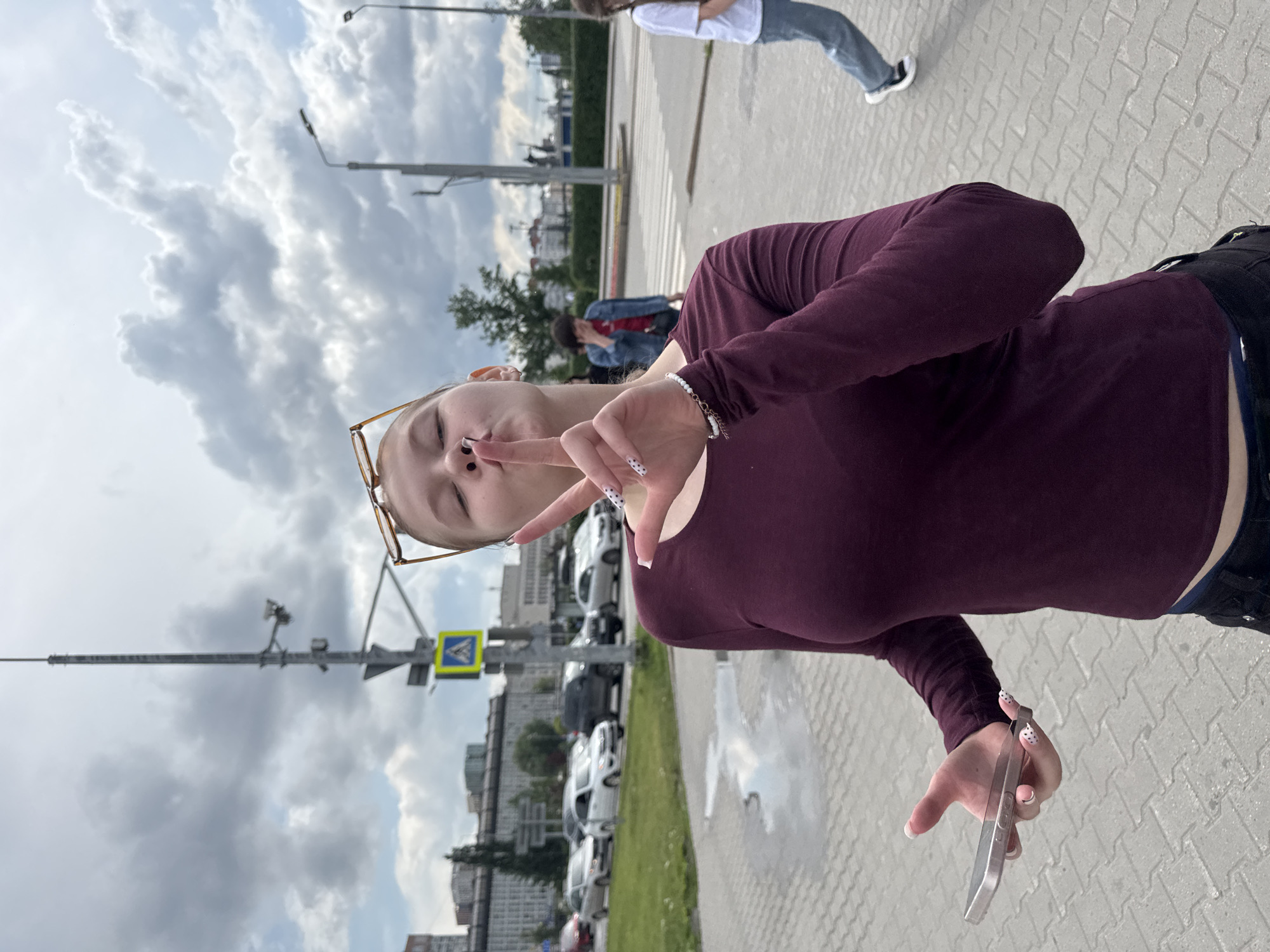 A young woman is the central subject of this street-level, full-body portrait, posing on a paved sidewalk in an urban setting in Perm, Russia. She is shown from the waist up, facing the camera with her eyes closed and lips pursed in a playful expression. Her right hand is raised, forming a peace sign, while her left hand holds a silver smartphone. She wears a long-sleeved burgundy top, dark trousers, and has square-framed glasses pushed up onto her head. A small nose piercing is visible, and her nails feature white polish, some with black polka dots.
In the mid-ground, another person wearing a white t-shirt and blue jeans walks away from the camera, and a third person in a denim jacket is partially visible further back. The background features a street with cars parked along the curb, multi-story buildings in the distance, and tall lamp posts and traffic light poles. A yellow square pedestrian crossing sign with a black walking figure is clearly visible. The ground is paved with grey blocks, and small puddles are scattered across the surface, indicating recent rain. The sky is heavily overcast with dramatic grey clouds, suggesting daytime during cloudy weather. The overall mood is casual and candid.