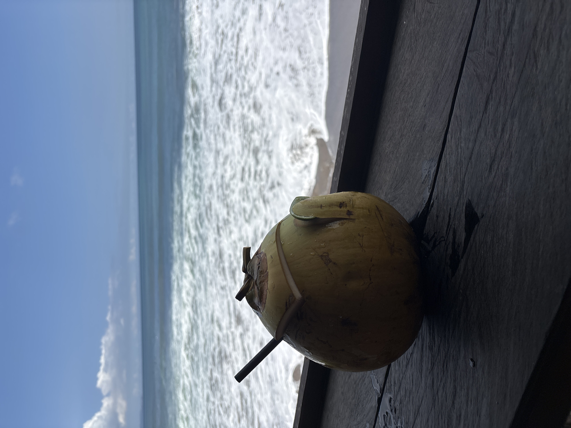 A coconut with a straw sits on a dark wooden surface, likely a table, in front of a beach scene. The beach features white waves crashing onto the shore, with the ocean stretching out to the horizon under a blue sky with white clouds. The image is likely taken in North Kuta, Indonesia.