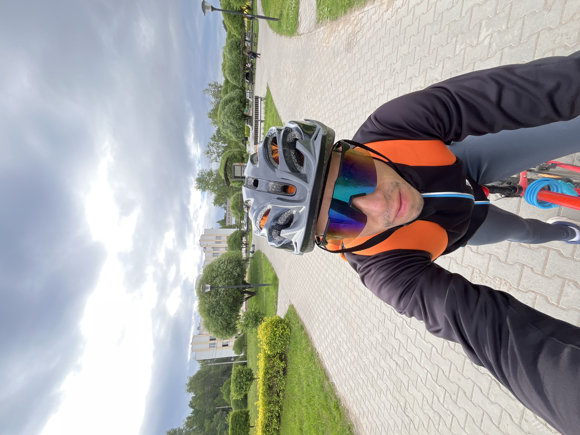 The image shows a person riding a bicycle on a paved path in Chaikovsky, Russia. The person is wearing a grey helmet, mirrored sunglasses, and a black long-sleeved shirt with an orange backpack. The path is made of interlocking concrete pavers and is surrounded by green grass and manicured bushes. In the background, there are trees, lampposts, and a light-colored building. The sky is cloudy.