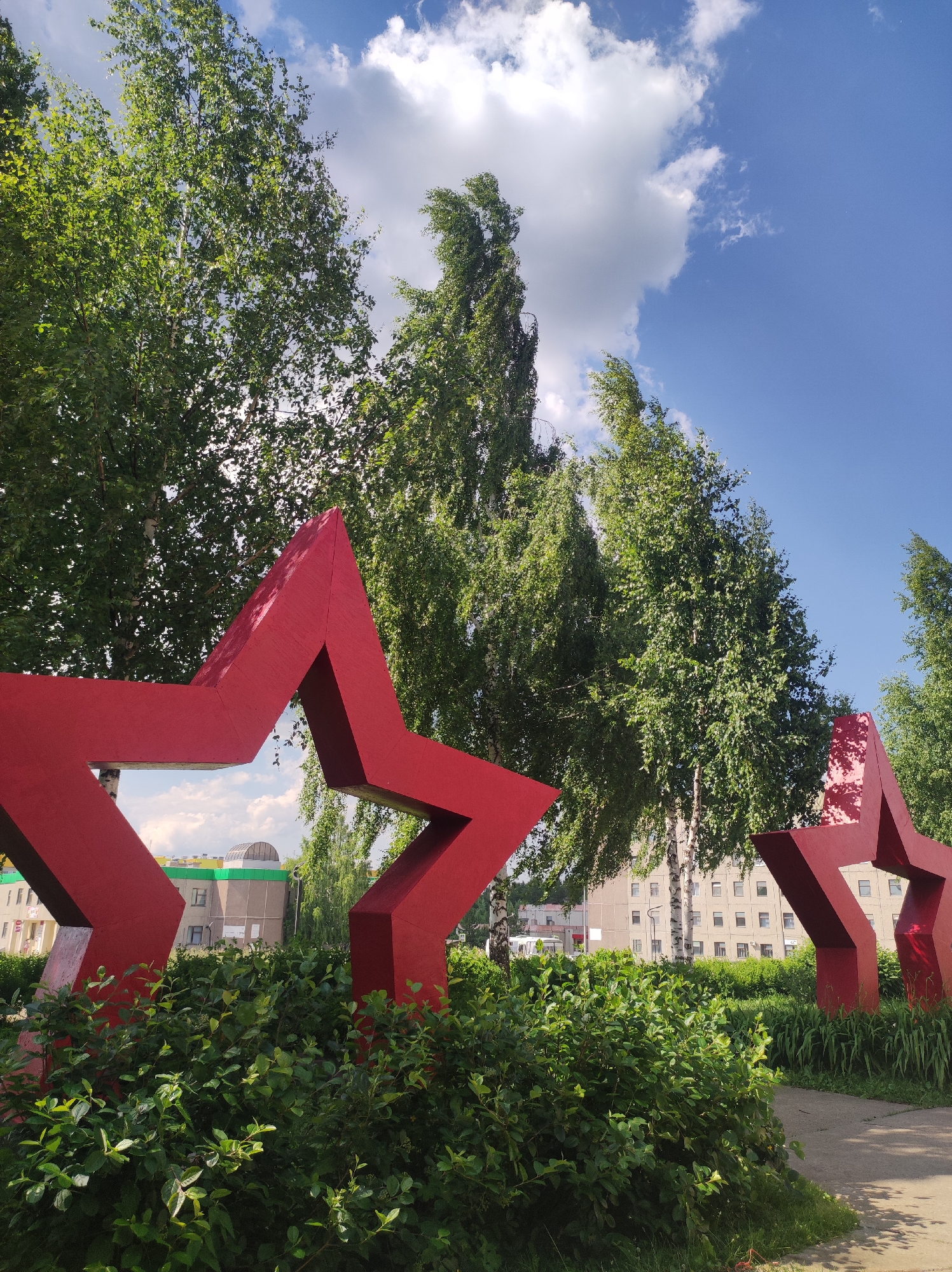 In the foreground, two large, bright red five-pointed stars, likely part of a monument or public art installation, are prominently displayed. The star on the left is positioned closer to the viewer and is slightly angled, allowing a glimpse of a building and a bright sky with clouds through its opening. The star on the right is further away and appears to be partially obscured by foliage. Lush green bushes and plants surround the base of these stars, creating a verdant landscape. Behind the stars, tall, leafy trees, primarily birches with their distinctive white bark, form a natural backdrop. Further in the distance, apartment buildings with multiple windows are visible, suggesting an urban or suburban setting. A paved pathway or walkway is visible to the right of the stars, leading into the scene. The sky above is a clear, bright blue with scattered white clouds. This scene is located in the city of Марковский, Russia. There are no people visible in the image.