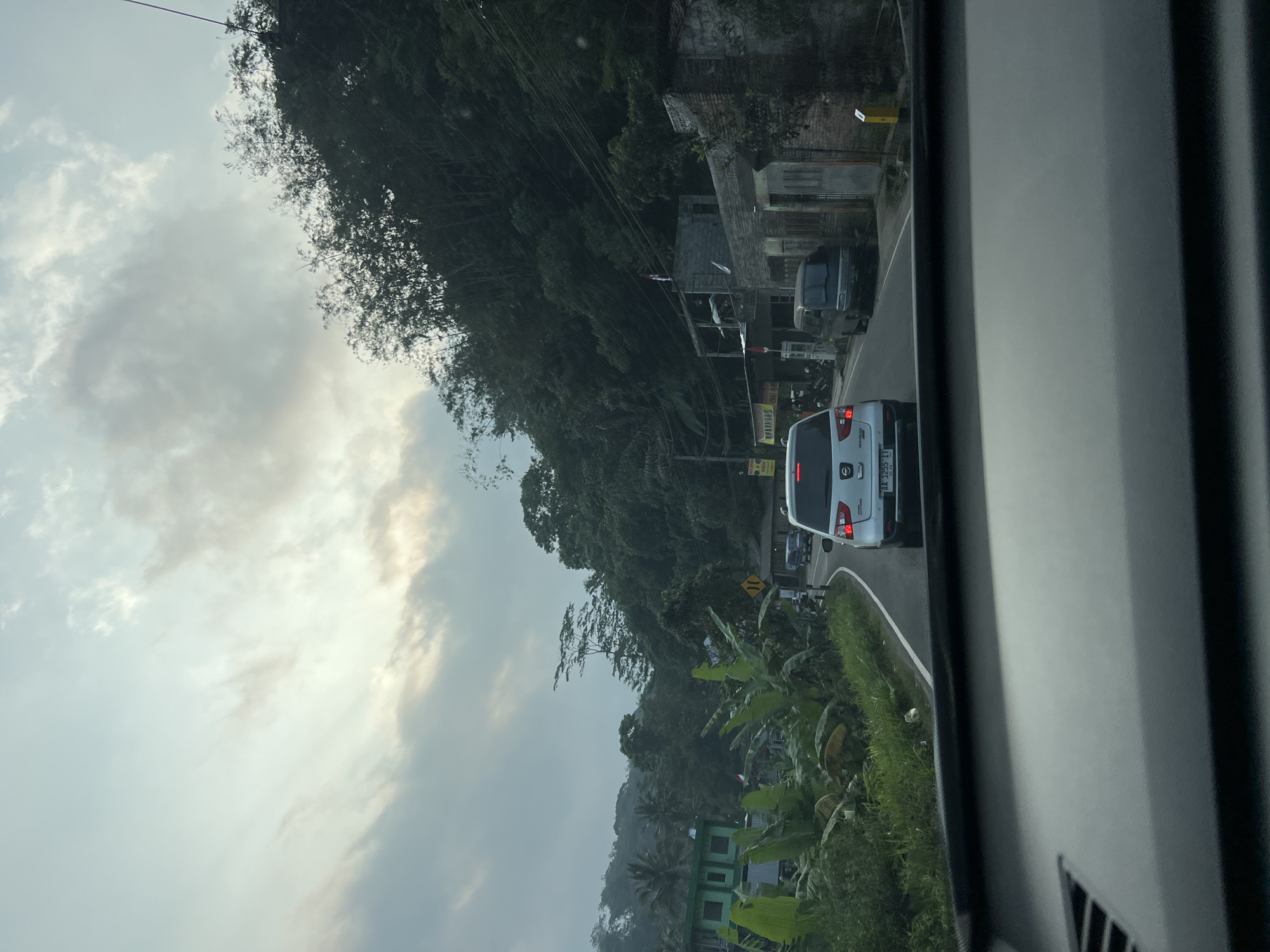The image shows a view from inside a vehicle traveling on a road in Beseran, Indonesia. The road is winding through a lush, green, hilly area with dense vegetation and trees lining the sides. The sky is overcast with soft, diffused light, suggesting it is either early morning or late afternoon. Several vehicles are visible on the road, including a white SUV in front, a grey van, and other cars further down. On the left side of the road, there are buildings, including a green structure with multiple windows and some houses. Banana plants and other tropical foliage are abundant. On the right side, there are more buildings constructed from brick and some shops. Traffic appears to be moving slowly. The overall scene depicts a typical rural or semi-urban environment in Indonesia, characterized by its natural landscape and roadside development.