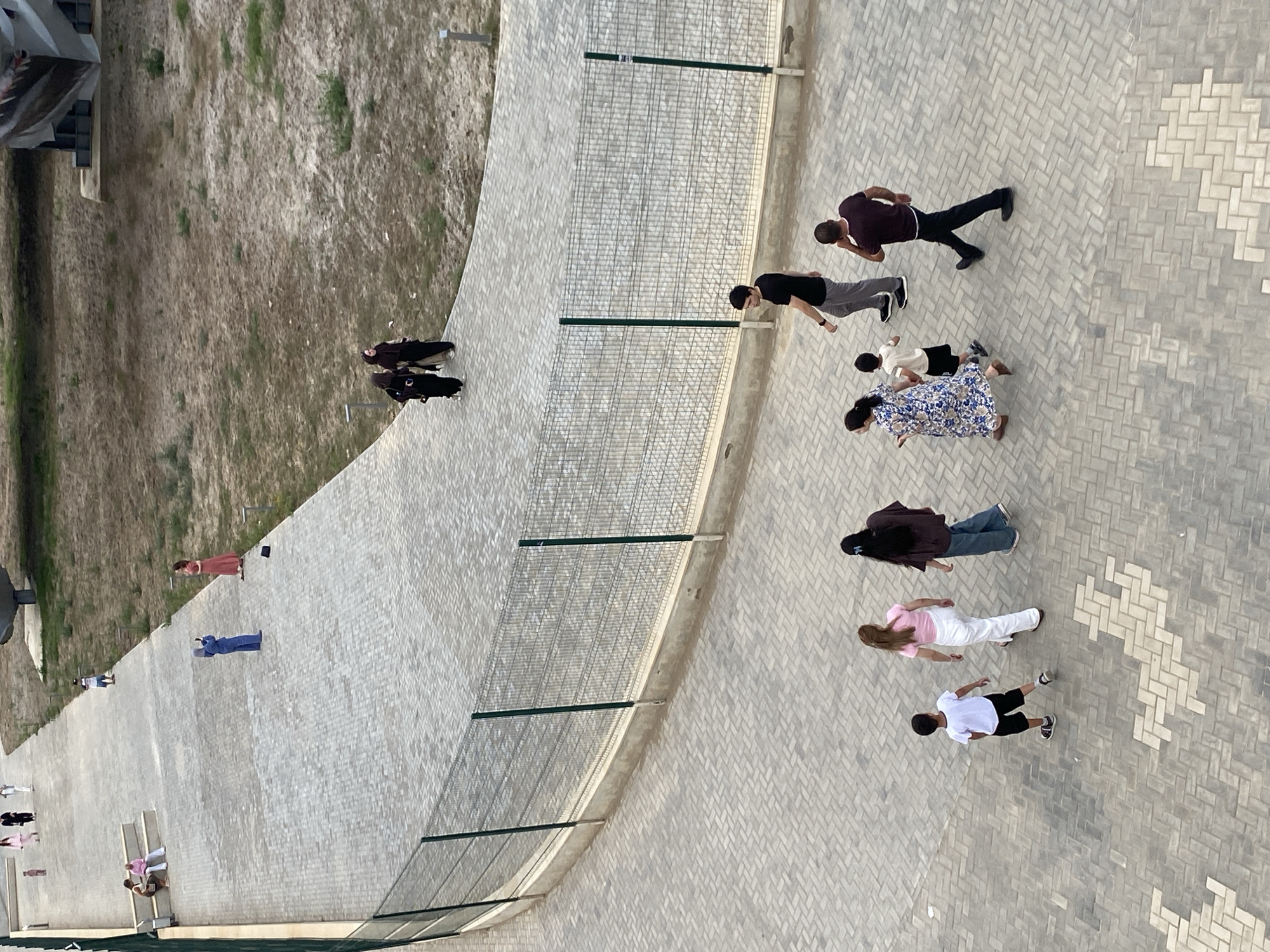 An aerial view captures a paved pathway in a park-like setting, possibly in Arablyar, Russia. A chain-link fence runs along the left side of the pathway, bordering a grassy, dry area. Several individuals are present. In the foreground, a group of three people, a woman in a floral dress, a young boy in a white t-shirt and black shorts, and a man in a dark t-shirt and black pants, are walking away from the camera. Behind them, a man in a black t-shirt and grey pants is walking to the right, and another man in a dark red t-shirt and black pants is walking to the left. Further up the pathway, two women in dark clothing are walking together. There is another woman in a pink top and white pants, and a person in a blue outfit standing and holding a phone. Two women are seated on steps on the left side of the pathway, and a young person is standing in the distance. The ground is covered in a patterned brickwork.