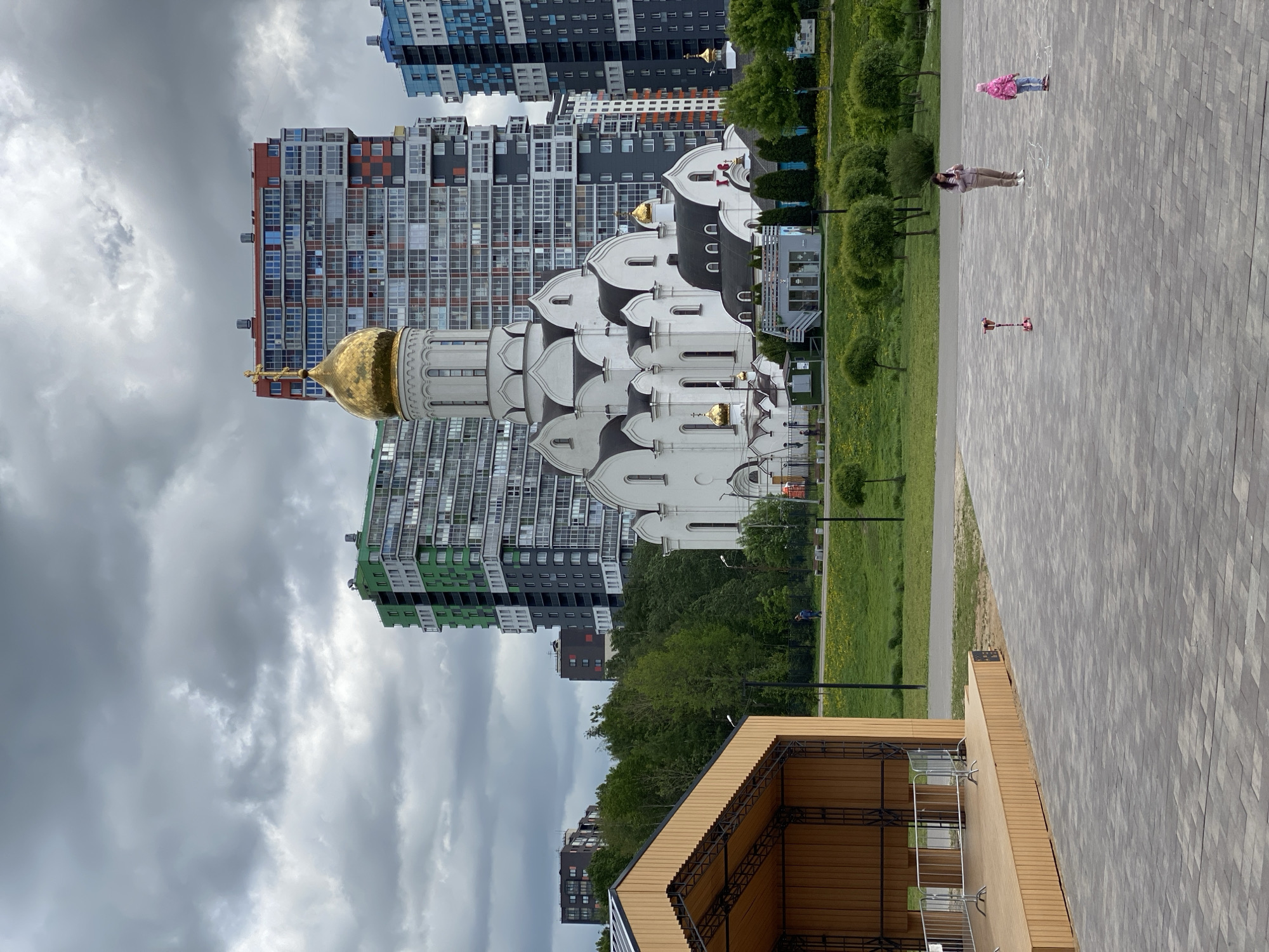 The image shows a modern urban landscape with a mix of architectural styles. In the foreground, there's a wooden structure, possibly a stage or pavilion. Adjacent to it is a paved area with two people standing and a scooter lying on the ground. Beyond the paved area, there is a green space with trees and a white church with golden domes. In the background, tall modern apartment buildings of various colors are visible, under a cloudy sky. The location is in the city of Unknown, Unknown.