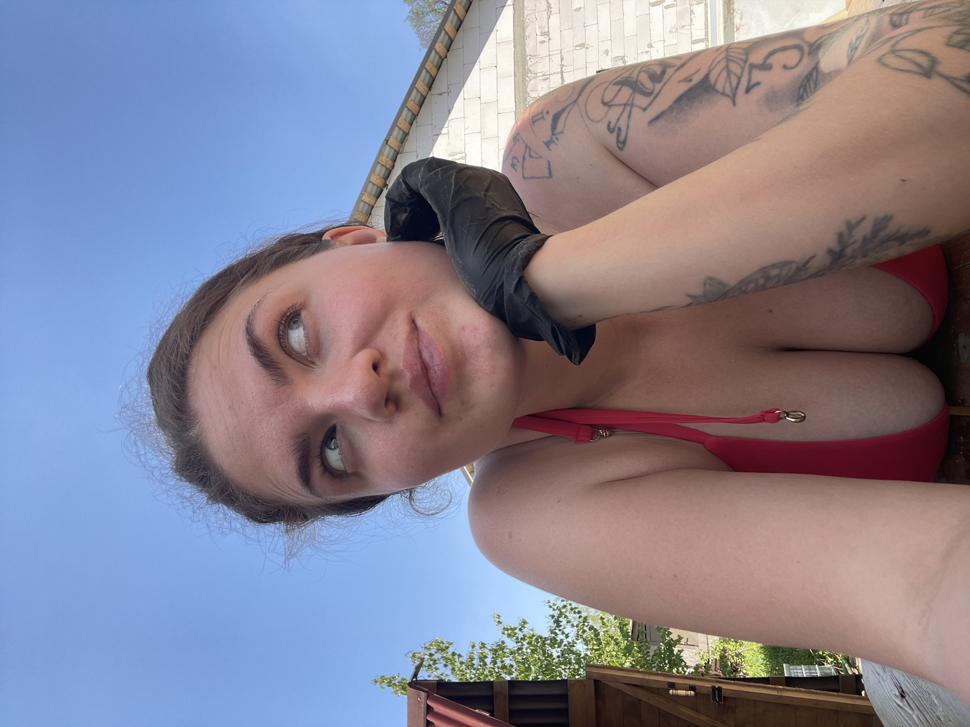 A person wearing a red swimsuit and black gloves is holding the ear of another person with a tattoo in an outdoor setting. The sky is visible in the background along with some foliage and a building made of white blocks. The location may be in the city of Мосметаллоконструкция Онт, Russia.