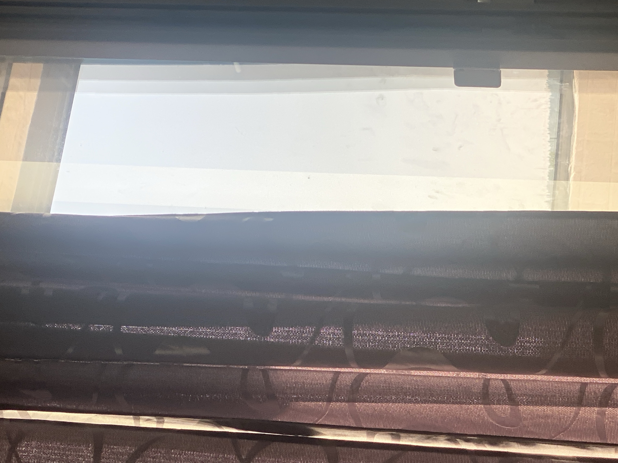 The image depicts a view through a window, partially obscured by draped curtains. On the left, a dark, patterned curtain with a subtle swirling design is visible, catching the sunlight and revealing its texture. To the right of the curtain, a window offers a bright view of an outdoor scene, dominated by a hazy, washed-out sky. A sliver of what appears to be the ground or a distant landscape with a horizontal line of trees or structures is discernible at the bottom of the window. A dark, rectangular object, possibly a door handle or a latch, is attached to the right side of the window frame. The lighting suggests it is daytime, with sunlight streaming in from outside. The scene offers no specific indicators to pinpoint its location in Novosibirsk, Russia.