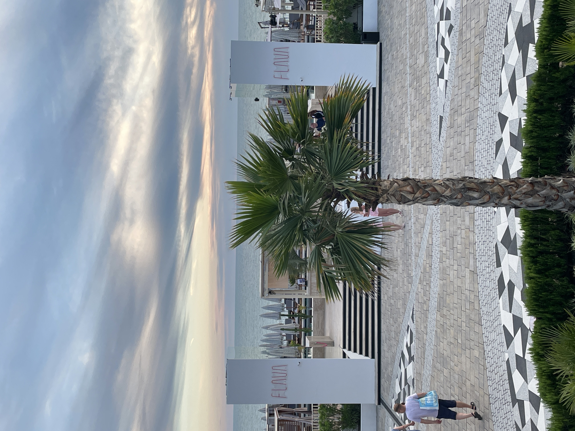 The image depicts a beachfront resort in Sochi, Russia, likely during the late afternoon or early evening. A prominent feature is a tall, slender palm tree in the foreground. In the background, two large white pillars are visible, each displaying the word "FLAVA" in red neon lettering. Beyond the pillars, there is a view of the sea under a cloudy sky with streaks of pink and orange from the setting sun. The resort area features rows of lounge chairs and umbrellas, suggesting a beach club or hotel setting. A man in a light-colored t-shirt and dark shorts, carrying a blue plastic bag, walks away from the camera on a patterned paved walkway. Other individuals are visible in the middle ground, including two women in pink attire near the steps leading up to the resort. The overall atmosphere is relaxed and suggests a vacation destination.
