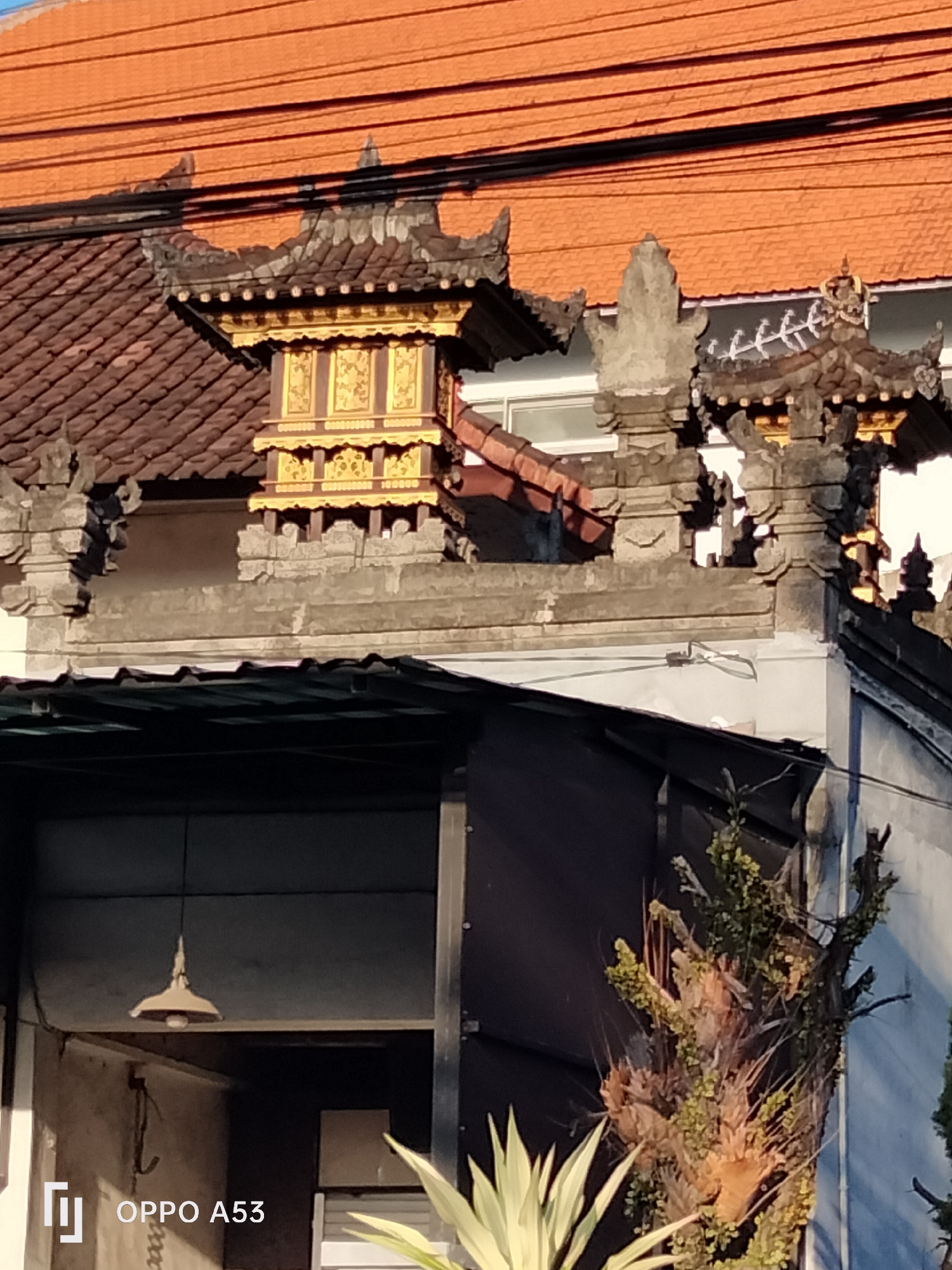 The image shows the rooftop of a building with traditional Balinese architecture. The roof is made of reddish-brown tiles, and there are ornate stone carvings and a golden structure with intricate details. A light fixture hangs from a dark ceiling below. In the foreground, there are plants, and a portion of a wall is visible. The Oppo A53 logo is visible in the bottom left corner. The scene appears to be in Denpasar, Indonesia, given the architectural style.