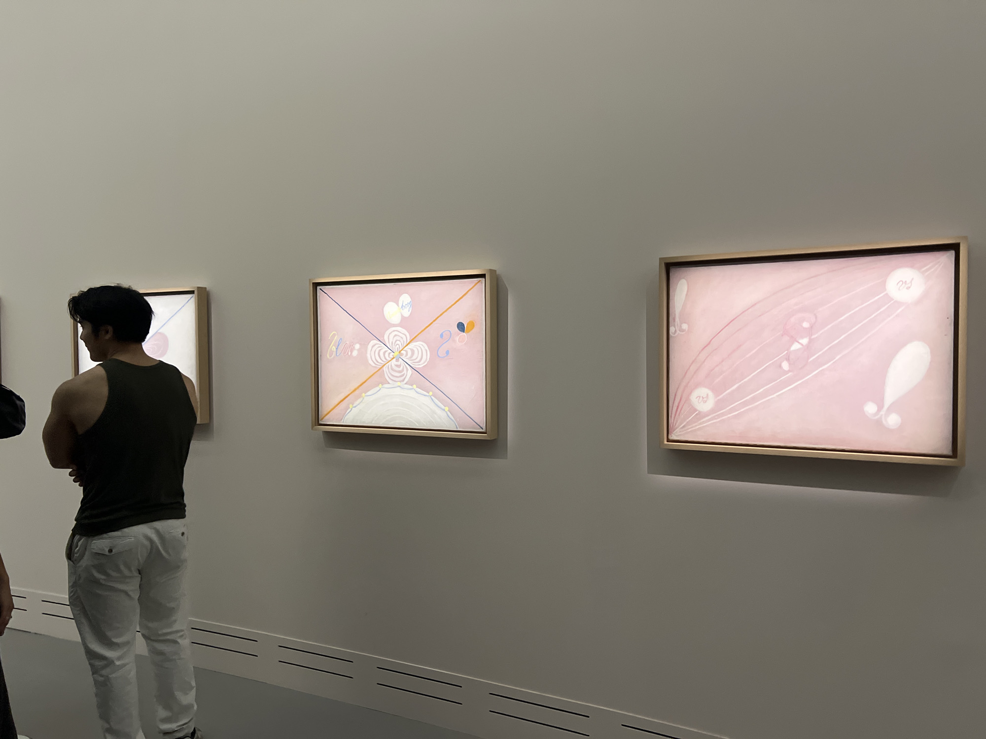 This is an eye-level, medium shot of a person standing in a museum gallery, facing away from the camera, looking at three framed abstract paintings displayed on a plain white wall. The person is wearing a dark sleeveless shirt and light-colored pants. The paintings are displayed horizontally, with a golden frame each. The leftmost painting is partially visible, with a pink background and a white circular motif. The middle painting features a pink background with a geometric design of radiating lines and curved shapes in white and blue, with some small yellow and blue dots. The rightmost painting also has a pink background and is characterized by flowing white lines and organic shapes. The gallery space is minimalist, with a white wall and what appears to be a white baseboard with horizontal black lines. The lighting is soft, illuminating the paintings and the gallery wall. The location is identified as the Pompidou Centre in Paris, France, a renowned modern and contemporary art museum.