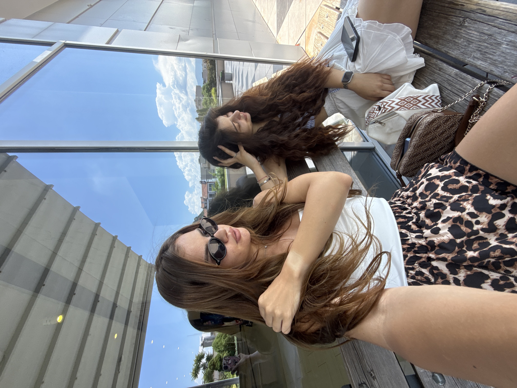 Two young women are sitting on a wooden bench outdoors, with a large glass window behind them reflecting the sky and surroundings. The woman on the left is in the foreground, facing to the right, wearing sunglasses, a white strapless top, and leopard-print shorts. She has long brown hair and is resting her left arm on her shoulder. The woman on the right is sitting next to her, facing left, with her right hand touching her hair. She has long, wavy brown hair and is wearing a white skirt and a top with blue stripes. A patterned handbag and a white tote bag are on the bench between them, along with a smartphone. The bench appears to be made of weathered wood. The reflection in the window shows a bright blue sky with white clouds, trees, buildings, and some street furniture, including a trash can. The overall environment suggests an outdoor seating area in a modern setting, possibly part of a building complex or a public space in the city of Plovdiv, Bulgaria, given the architectural style and the sunny weather.