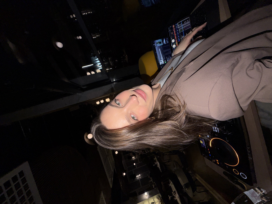 The image depicts a female subject situated at a DJ setup within the Terra Rooftop venue in Budva, Montenegro, at night. The subject, appearing to be in her 20s or 30s with long brown hair and light-colored eyes, is wearing a light blue top, a gold necklace, and a brown blazer. She is angled slightly to her right, gazing directly at the camera with a subtle smile.
In the foreground, to the subject's left (viewer's right), a DJ controller or turntable with illuminated elements is visible on a booth, alongside a glass containing a clear liquid. To her right (viewer's left), two laptops are positioned. One laptop screen displays what appears to be DJ software with audio waveforms and track information; the other shows a music streaming application. The subject's right hand rests near the keyboard of the rightmost laptop.
The background reveals a dimly lit interior space, indicative of a modern restaurant or lounge, with various tables, chairs, and hanging light fixtures. A yellow accent chair is partially visible behind the DJ station. Beyond a large window, city lights are visible against the dark night sky, with a clearly illuminated "TERRA" sign confirming the venue's name.