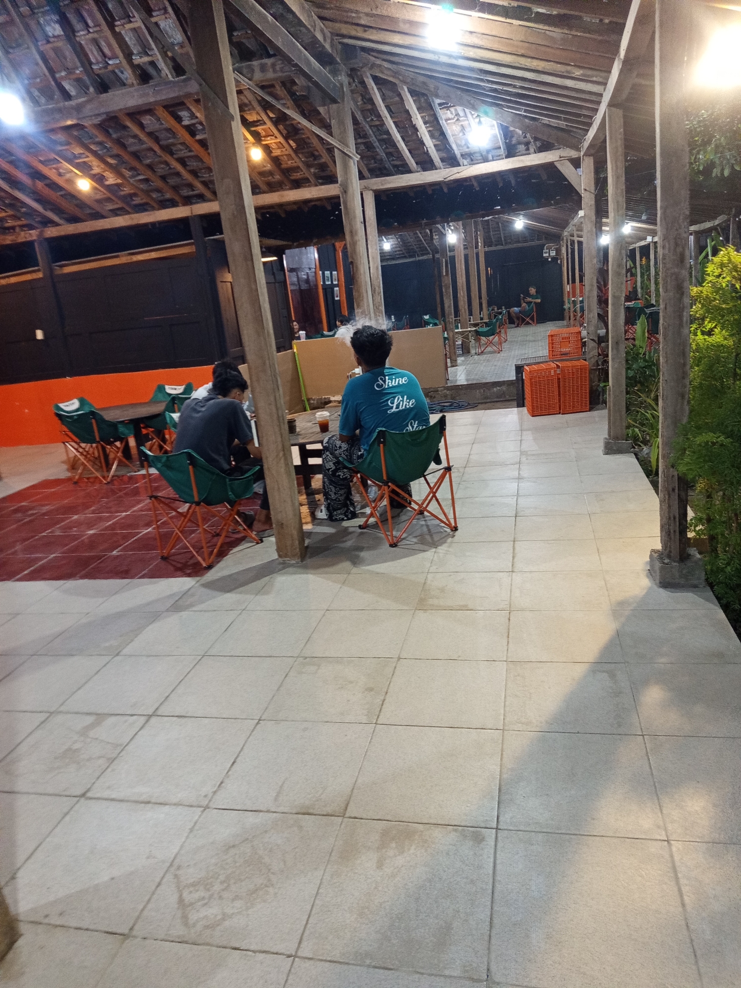 The image shows an outdoor seating area at what appears to be a cafe or restaurant in Becirongengor, Indonesia. The structure features a rustic wooden roof supported by thick wooden posts. The floor is paved with large, light-colored square tiles. In the foreground, a pathway with tiled flooring is visible, with shadows cast by overhead structures and possibly trees.
There are two individuals seated at tables. One person, wearing a dark t-shirt, is seated at a table on the left side of the image, facing away from the camera. They are sitting in a foldable green chair with an orange frame. Another person, wearing a blue t-shirt with the text "Shine Like Star" printed on the back, is seated at a table on the right, also facing away from the camera and sitting in a similar green and orange foldable chair. This person is holding a beverage in a clear cup.
Various other elements are present in the environment, including more foldable chairs, tables, orange crates, and some greenery. The lighting suggests it is either late afternoon or evening. The overall impression is a casual, open-air dining or gathering space.