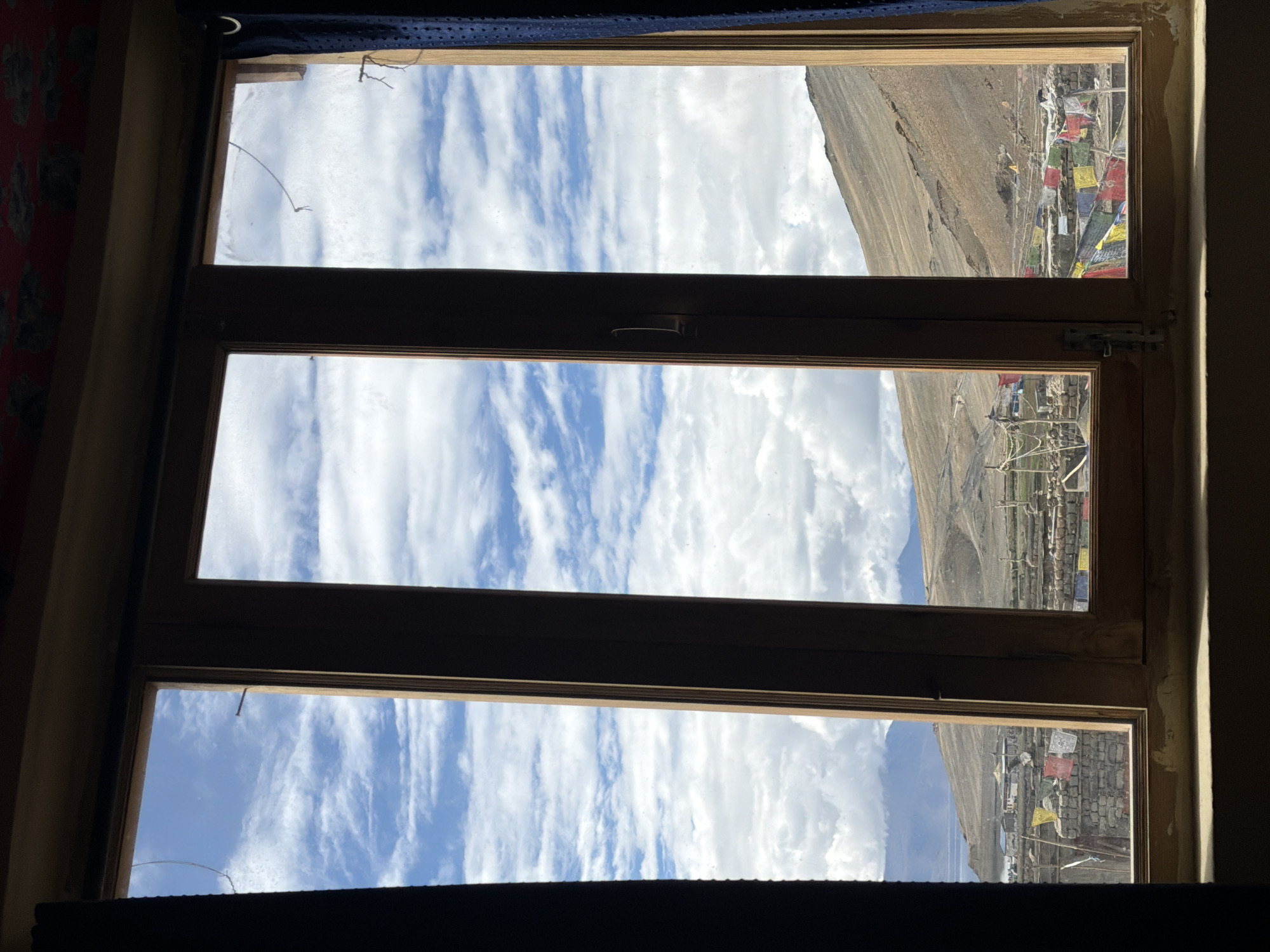 The image shows a view through a wooden window frame, divided into three vertical panes, looking out onto the village of Karzok, India. The sky is filled with white, puffy clouds against a bright blue expanse. In the middle and right panes, the landscape slopes upwards, revealing barren, brown hillsides with visible textures.
The village appears to be nestled at the base of these hills. Buildings with stone walls and flat roofs are visible. Prayer flags, colorful and rectangular, are strung across several areas, fluttering in the breeze. These flags suggest a spiritual or cultural significance to the location. The overall environment appears dry and mountainous, characteristic of high-altitude regions. The window frame itself shows some wear and tear, with a metal latch on the central pane. The foreground, where the windowsill is located, is dark and indistinct.