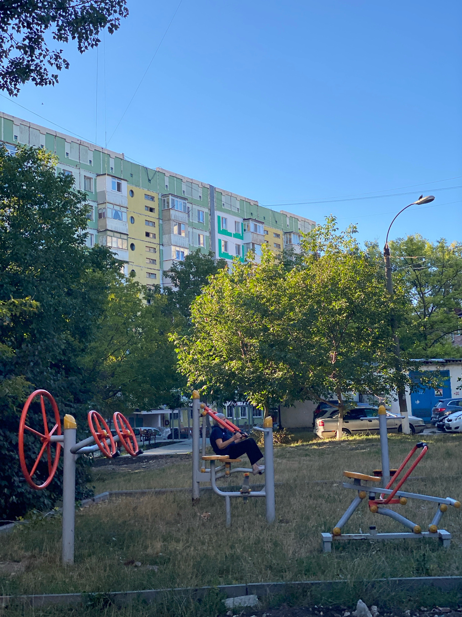 The image depicts an outdoor exercise equipment area located in front of a Soviet-era apartment building in the city of Chișinău, Moldova. The building, which appears to be in a state of disrepair, is multi-story with a colorful facade featuring green and yellow paint. Several trees with lush green foliage are scattered throughout the scene, casting shadows on the ground. The exercise equipment includes a stationary bike with large orange wheels and a rowing machine. A person is seated on the stationary bike, dressed in dark clothing and appearing to be engaged in a workout. The ground is covered in dry grass, and a paved walkway runs along the foreground. Several cars are parked in the background, and a street lamp stands to the right of the frame. The sky is clear and blue, suggesting a sunny day.