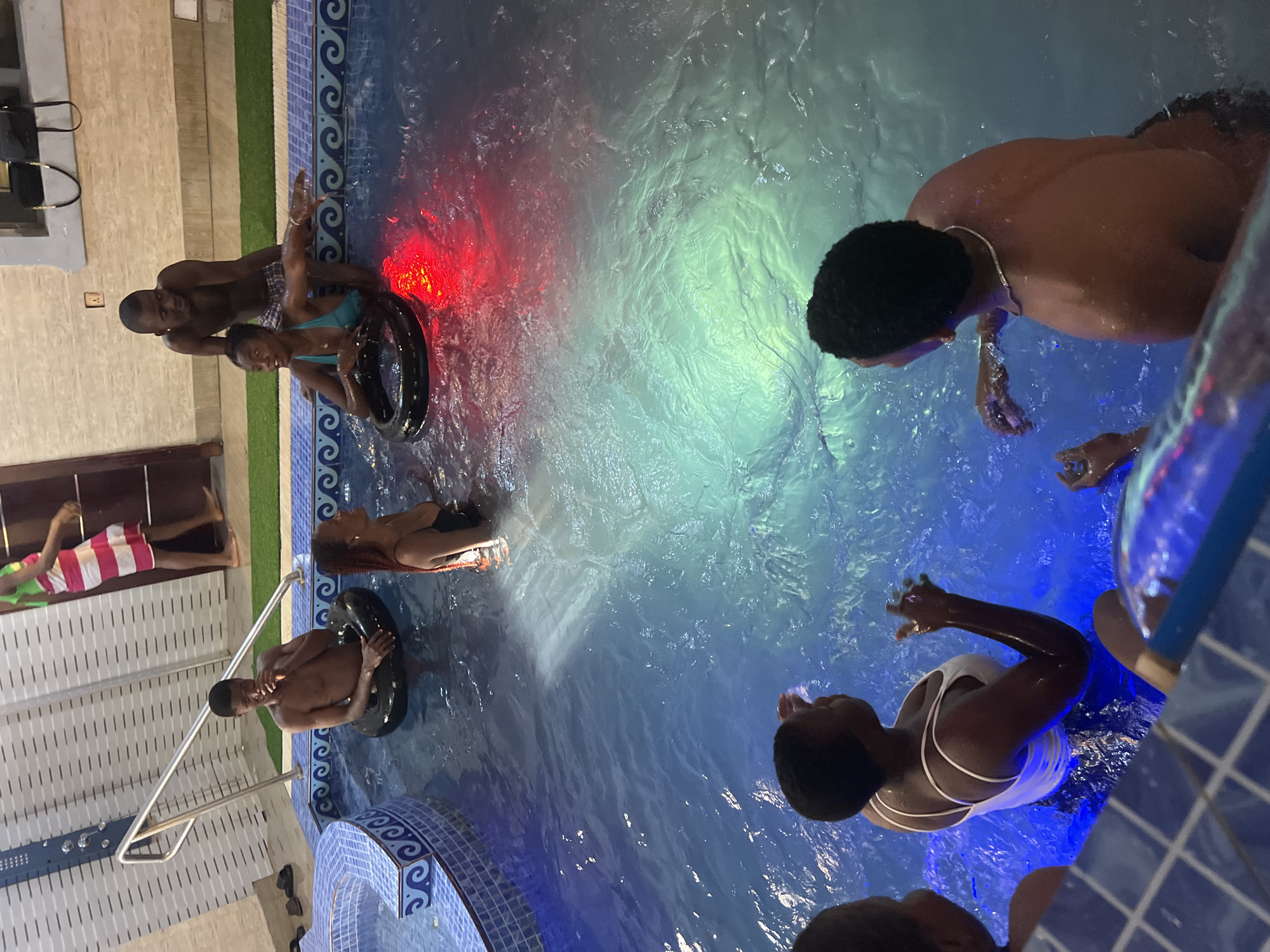 The image depicts a group of people in a swimming pool at night. There are approximately 6 people visible, some in inner tubes, others standing in the water. The pool is illuminated with blue and red lights. Some individuals are wearing swimwear. The surrounding area suggests an indoor or enclosed pool setting with tiled walls. Location is Unknown, Unknown.