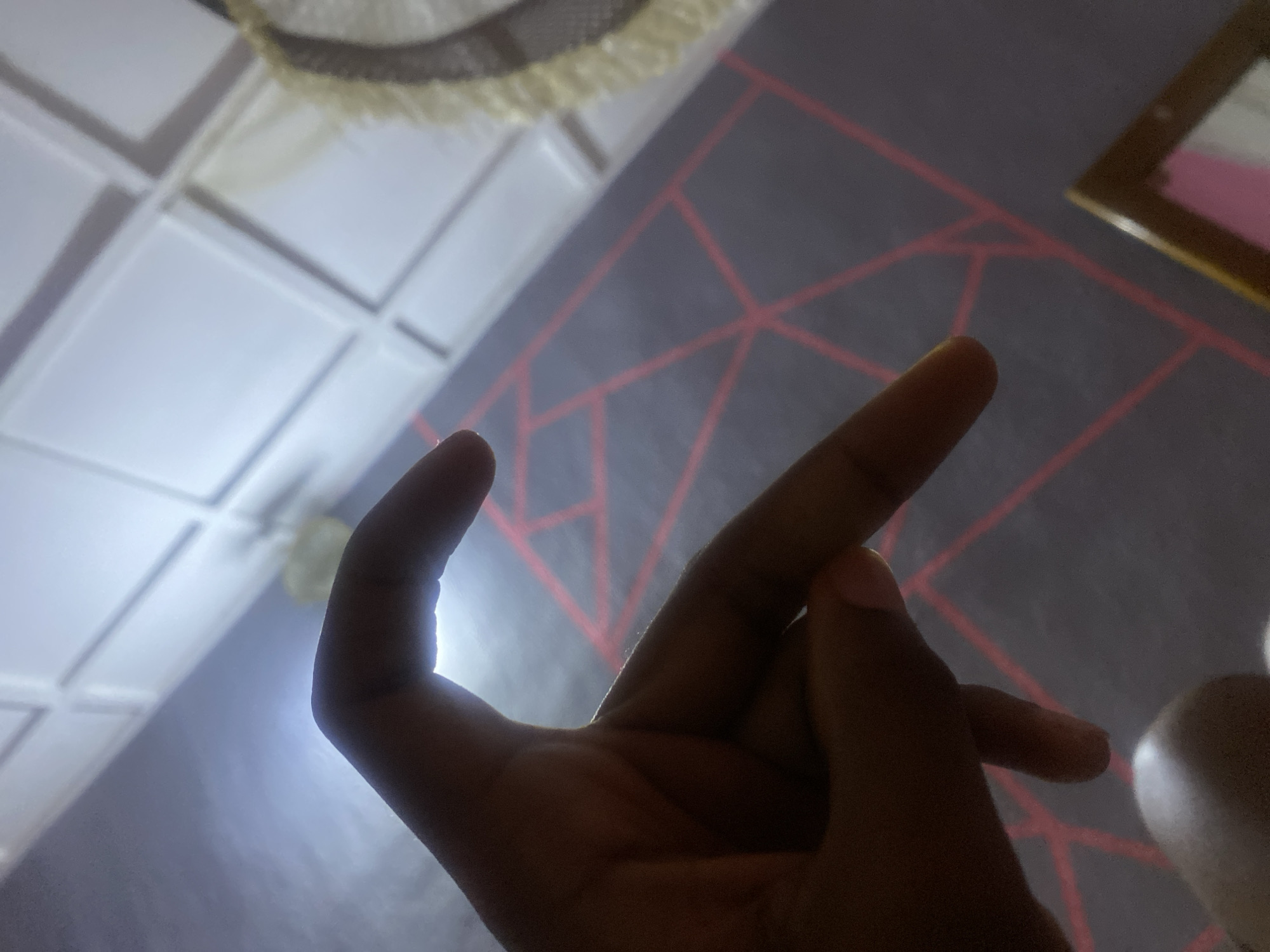 The image displays a human hand, primarily in silhouette, positioned in the foreground against a strongly backlit indoor environment identified as being in the city of Jos, Nigeria. The hand is posed with the thumb and fingers curved, appearing dark due to the bright light source originating from behind it. The background features a structured ceiling composed of white, rectangular paneled sections in the upper portion of the frame. Below this, a wall with a dark grey or purple tone is visible, adorned with a repeating geometric pattern of red or pink lines forming interconnected triangles and other polygonal shapes. In the upper right quadrant of the background, a blurry, light-colored object with a textured or fringed perimeter is partially discernible. The lower right corner of the image reveals a segment of a rectangular object with a light brown frame, possibly a mirror or a picture frame, reflecting colors that appear pink and white.
