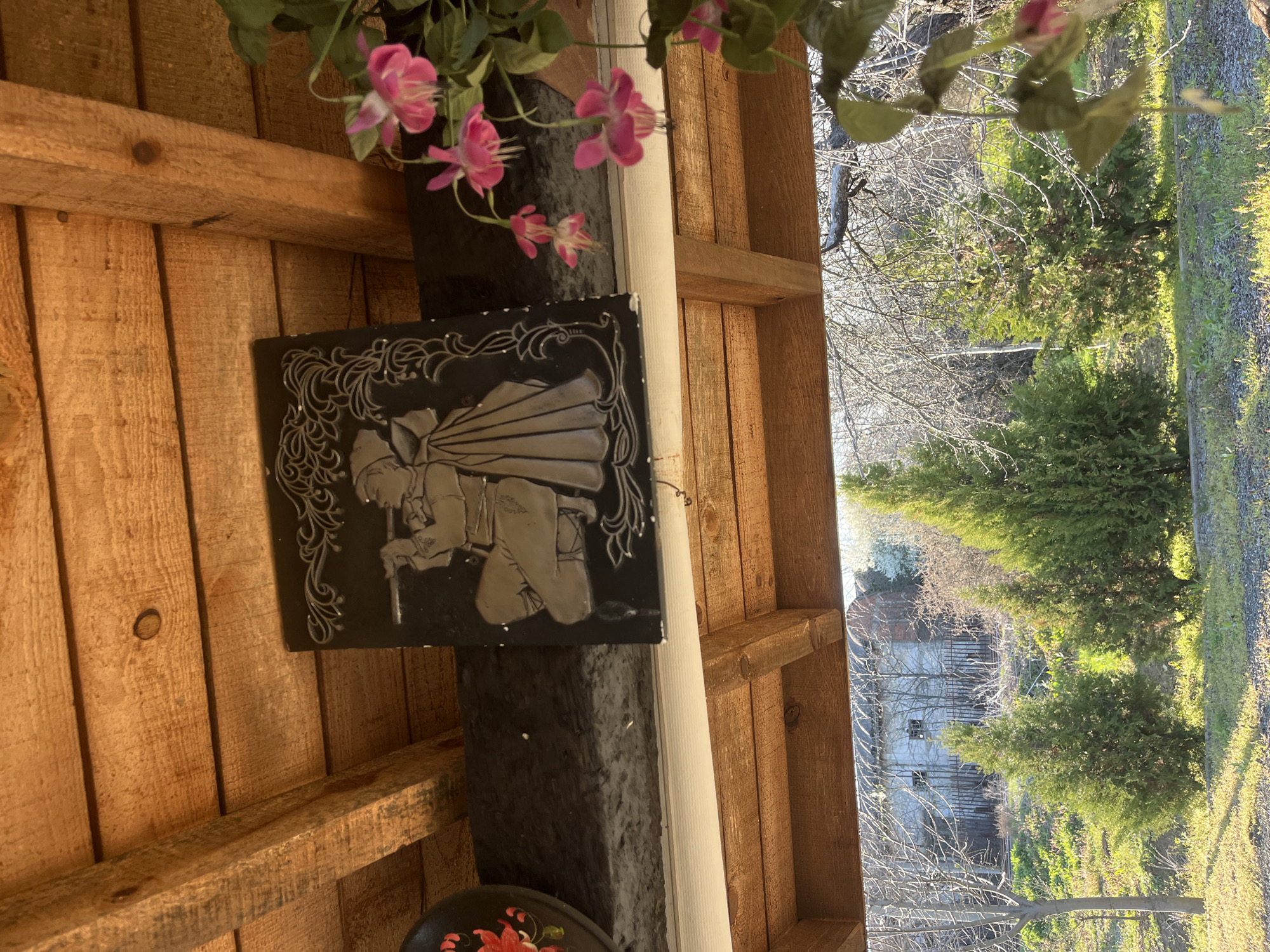The scene depicts a partially covered outdoor space, potentially a porch or veranda, in Lozarevo, Bulgaria. The dominant materials appear to be wood, comprising the structure’s ceiling and walls. A decorative black square plaque adorned with a silver-toned figure of a bent-over person holding a staff is prominently displayed. Artificial pink flowers add a touch of color to the scene. Through the aperture of the covered area, the image reveals a natural environment including trees and vegetation; a weathered building is visible in the background.