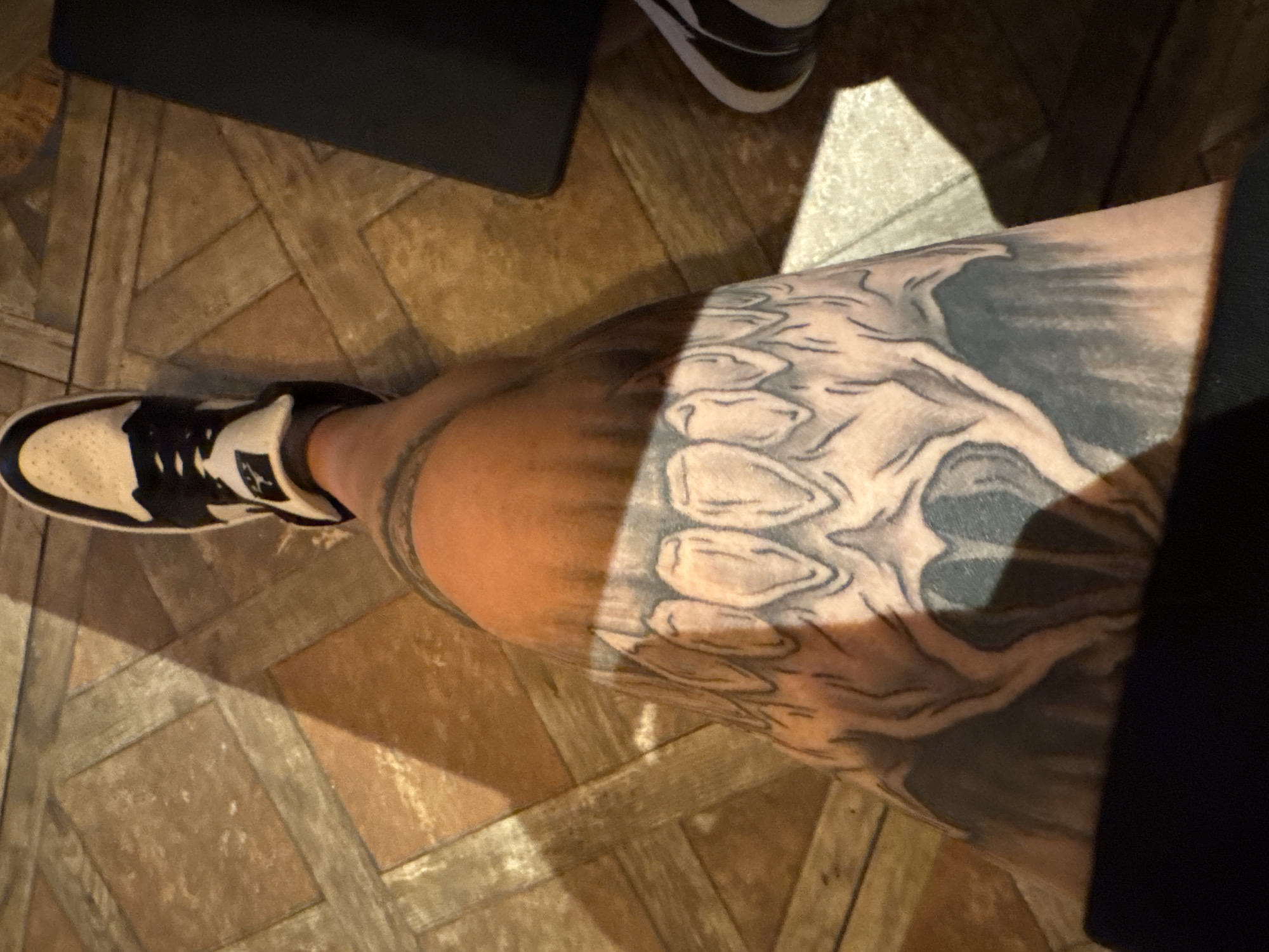 The image shows a close-up view of a person's leg with a large tattoo of a skull's teeth. The person is wearing a black and white sneaker. The leg and shoe are positioned over a tiled floor with a diamond pattern, which appears to be in a dimly lit indoor environment. Sunlight is casting a strong shadow across the floor and the person's leg. No specific location in Varna, Bulgaria can be determined from the visual information provided.