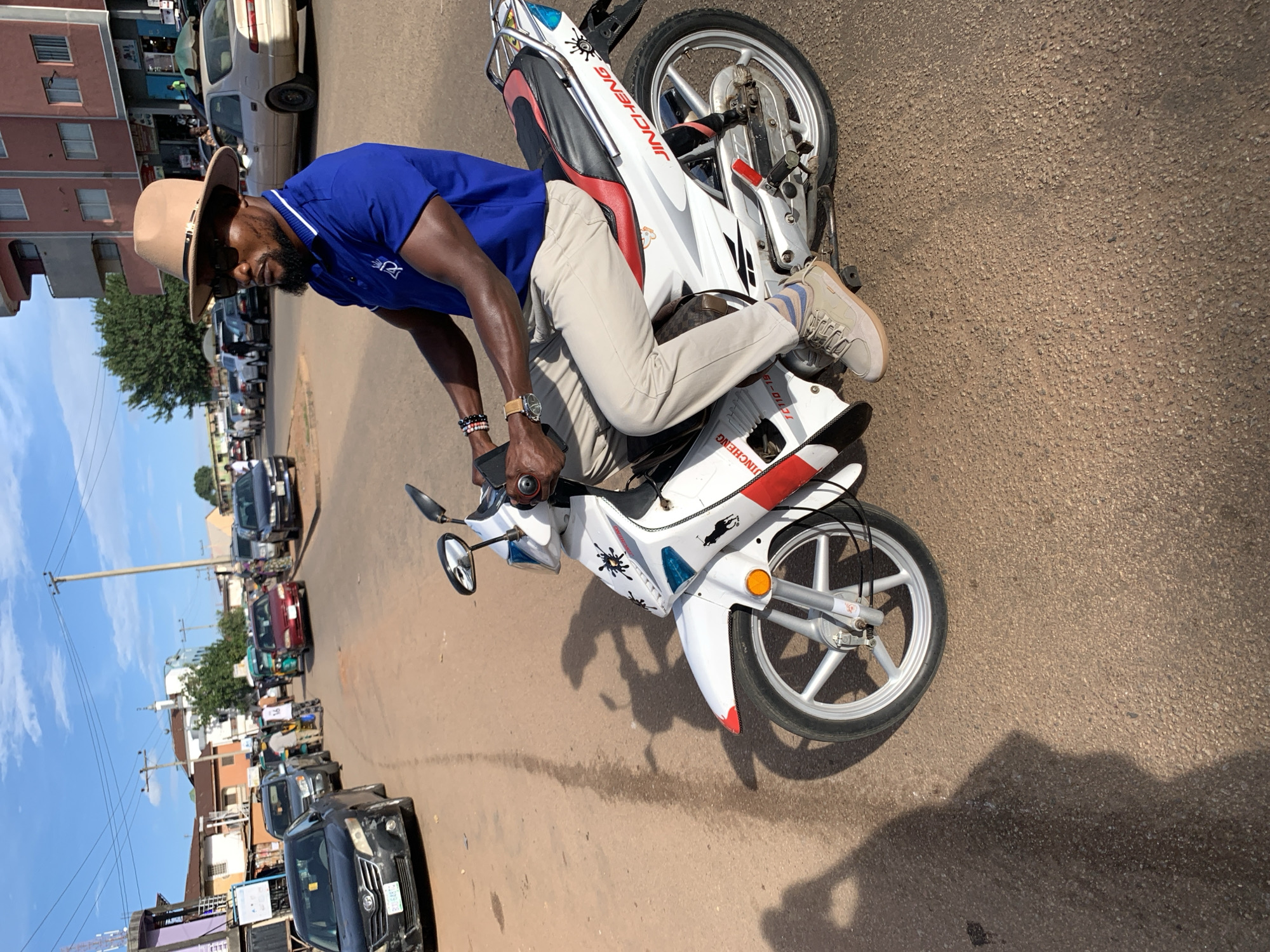 A man wearing a brown fedora hat, sunglasses, a blue polo shirt, and light-colored pants rides a white motorcycle with red and black accents down a paved road in Jos, Nigeria. The man is looking to his left, and his hands are on the handlebars. The motorcycle has a visible brand name that appears to be "JINCHENG" on the side. In the background, there are buildings, trees, and other vehicles, including cars and what appears to be a tuk-tuk. The sky is blue with some clouds. The lighting suggests it is daytime, possibly late afternoon due to the shadows. The road surface is asphalt with some patches of dirt or debris.