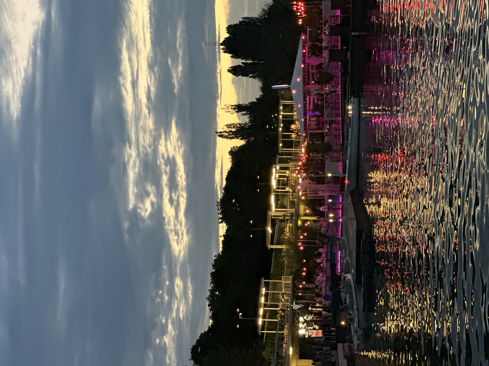 The image depicts a lively outdoor dining area on the Danube Canal in Vienna, Austria, at dusk. Numerous patrons are seated at tables, illuminated by string lights and the warm glow of the setting sun. The scene is set against a backdrop of lush green trees and a dramatic sky filled with textured clouds, with hints of yellow and orange near the horizon. Reflections of the lights and the sky dance on the water's surface, creating a vibrant and dynamic atmosphere. Several boats are docked along the canal, adding to the waterfront ambiance. The architecture of the dining area features open-air structures with canopies and railings, suggesting a modern and relaxed establishment.