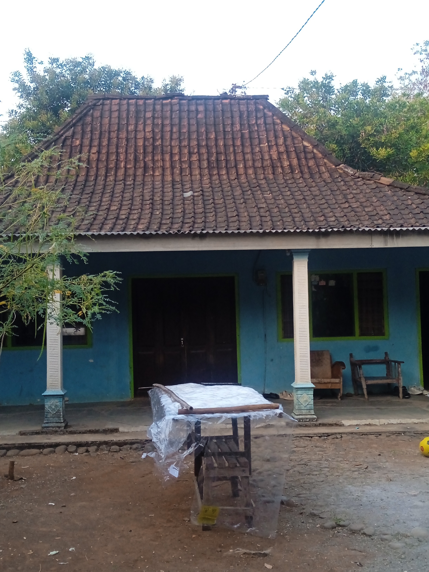 This image depicts a rustic house in Jatisrono, Indonesia. The house features a blue exterior with a prominent, tiled roof. Two wooden chairs, one upholstered and one a simple bench, are placed on the porch. In front of the house, a small table covered in plastic sheeting is visible, with a yellow ball resting on the ground to the right. The ground in front of the house is dirt, with scattered stones and vegetation. A power line runs overhead. The scene appears to be set in the late afternoon, with soft light casting shadows.