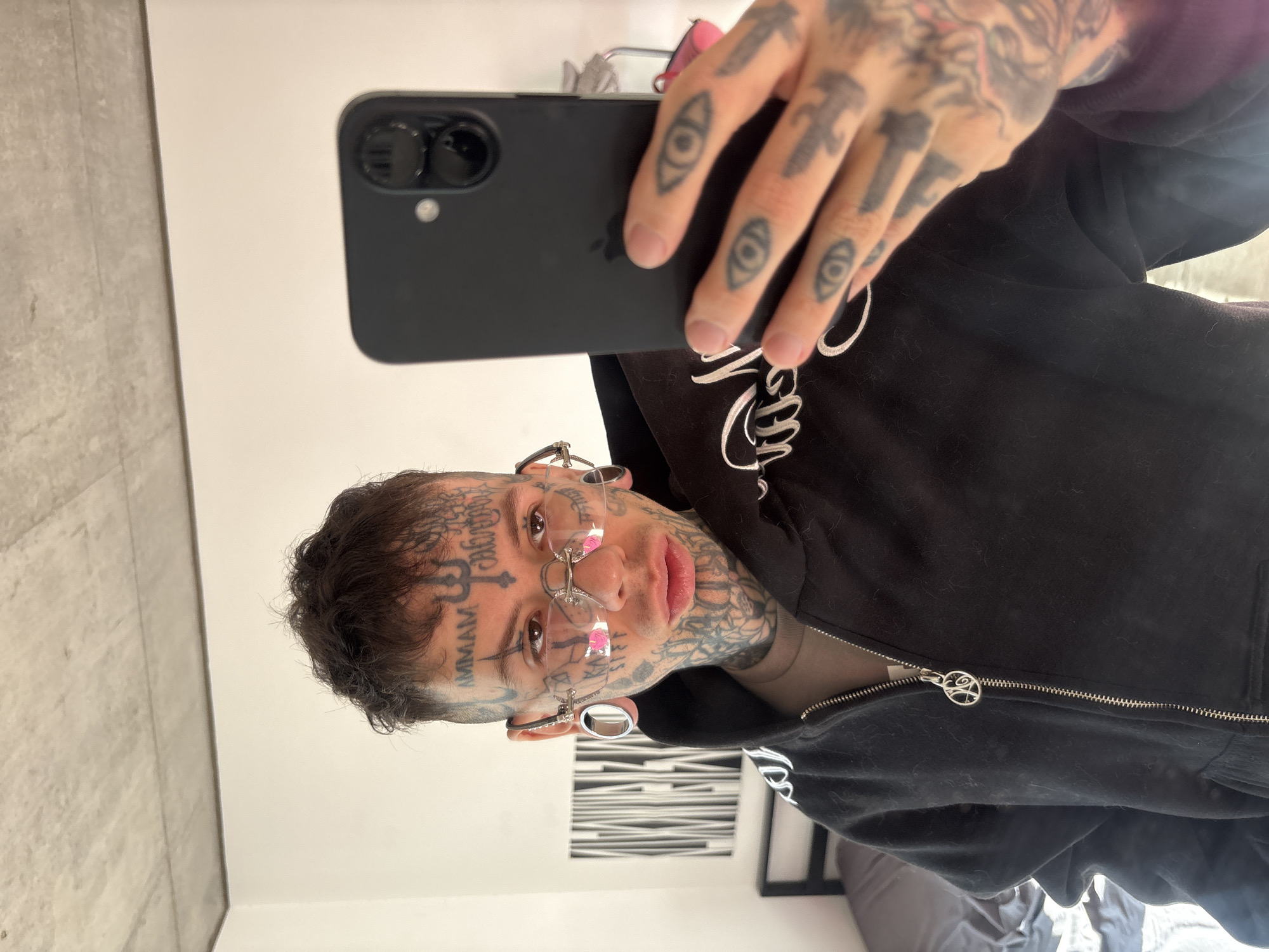 A selfie of a fair-skinned person with extensive tattoos on their face and hands. The person is wearing a black hooded jacket with a silver zipper. Their hair is short and dark. They have stretched earlobes with metal tunnels and clear framed glasses. The hand holding the phone has tattoos on the fingers. The background is a white wall with a textured gray section and a black and white artwork visible. The location cannot be determined from the image.