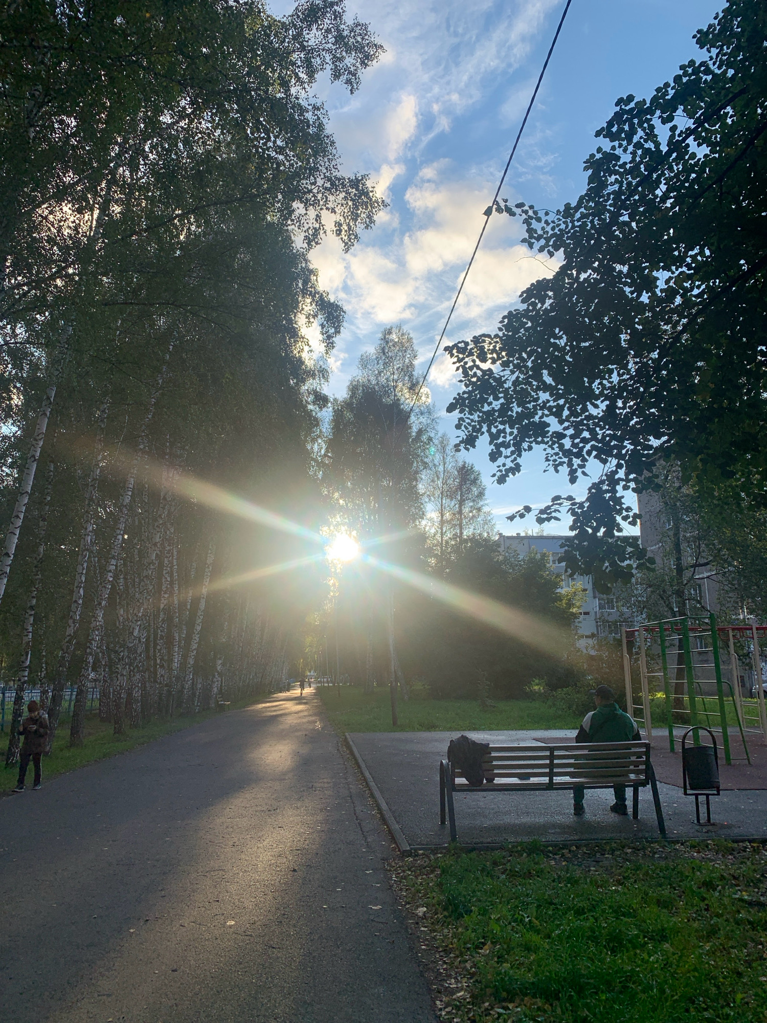 The image captures a serene outdoor scene on a sunny day, possibly in the city of Prokopyevsk, Russia. A paved pathway lined with birch trees creates a shaded walkway, with sunlight piercing through the leaves, casting dappled shadows and lens flares across the scene. In the distance, a person is seen cycling along the path.
To the right of the pathway, a park bench is occupied by an individual wearing a green tracksuit, facing away from the camera. A bag rests on the bench beside them. Adjacent to the bench is a trash can. Further to the right, a playground structure with green and silver elements is visible. Beyond the trees and playground, a multi-story residential building can be seen. The sky is a clear blue, dotted with scattered white clouds. The ground is covered with grass and fallen leaves, suggesting it may be autumn.