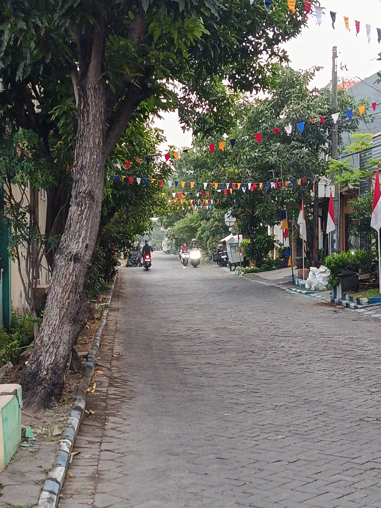 This image depicts a street scene in the city of Keboansikep, Indonesia, possibly during a celebration given the numerous small, colorful flags strung across the street and along the buildings. The road is paved with cobblestones and lined with trees and houses. Several motorbikes are present, with at least three individuals riding them. One person on a motorbike is in the foreground, moving away from the camera, while two others are further down the street. Red and white Indonesian flags are also visible, mounted on poles outside some of the buildings on the right side of the street. The overall lighting suggests it is daytime, perhaps late afternoon or early morning, with an overcast sky.