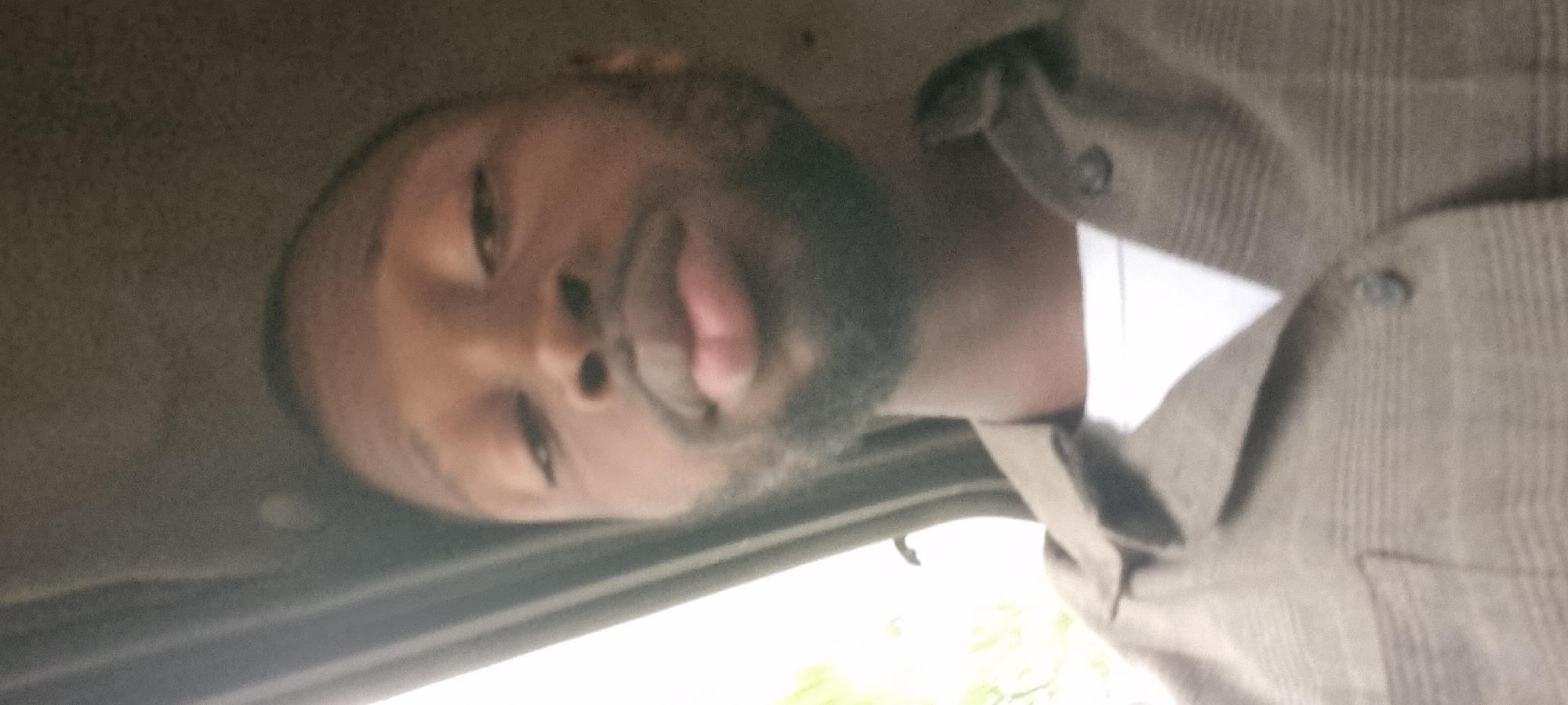 A close-up, indoor, selfie-style photograph shows a Black man with a light beard and a subtle smirk. He is wearing a brown collared shirt over a white t-shirt. The lighting is uneven, with the right side of his face and body being brighter than the left. The background is dark and indistinct, suggesting an interior space with possibly a window on the left side casting some light. The man's head is tilted, and his gaze is directed towards the camera. The image is slightly blurred, particularly around the edges. The location in Agege, Nigeria, is not discernible from the visual information provided in the image.