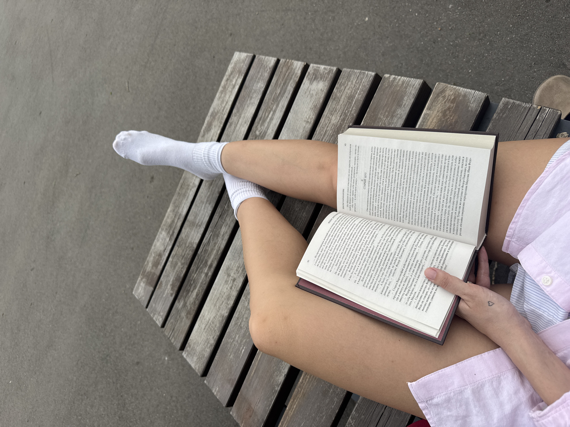 A high-angle, medium shot depicts a person sitting on a wooden bench, engrossed in reading a book. The bench, constructed from light-colored wooden planks, is set against a neutral gray concrete surface.
The person is seated with their legs crossed, wearing white socks. Their right hand is holding the open book. A heart tattoo is visible on their hand. The book's pages are filled with dense text in Cyrillic. They wear a pink and white striped button-down shirt.
The setting appears to be outdoors.