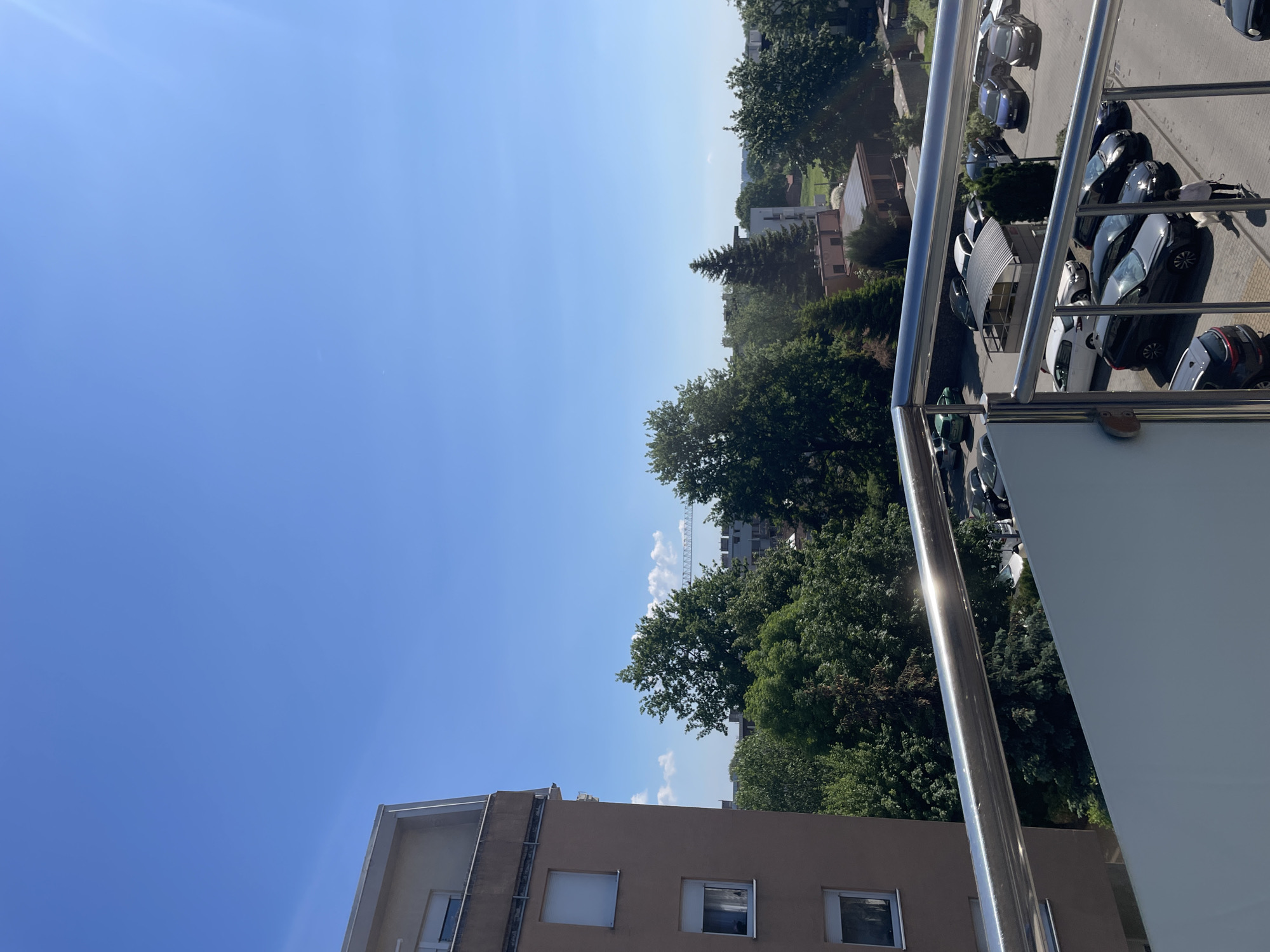 The image shows an outdoor scene from a high vantage point, likely a balcony. The sky is blue and mostly clear. A building with beige walls and windows is in the foreground. Beyond the building and extending towards the horizon are green trees and other buildings. To the right, a metal railing of a balcony is visible, along with a parking lot filled with various vehicles. The location is in an urban area, but the specific city is Unknown, Unknown.