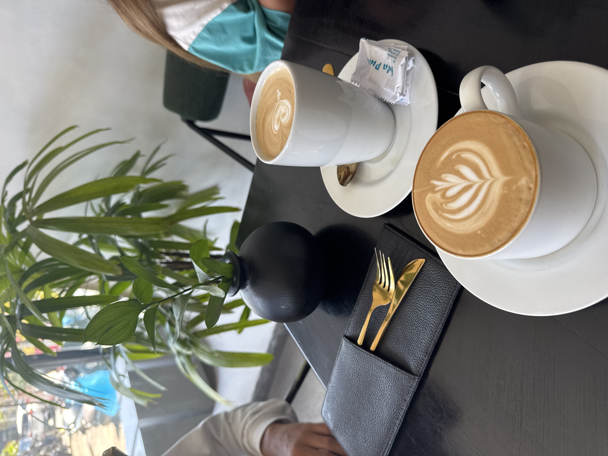 The image presents an overhead, angled view of a table in what appears to be an indoor cafe or restaurant setting. Two white ceramic coffee cups with matching saucers are prominently featured. Each cup contains a coffee beverage with intricate latte art; one displays a heart pattern, while the other shows a stylized leaf or fern design. On the saucer of the heart-patterned coffee, two small white sugar packets are visible, with partial text including "A la P" and "sugar".
To the left of the cups, a black, spherical vase holds several sprigs of green leafy plants, contributing to the decor. To the right, a black leather-like pouch contains gold-colored cutlery, consisting of a fork and a knife. The table itself is dark-colored.
Portions of two individuals are visible. In the upper left quadrant, the head and shoulder of a person with long brown hair, wearing a white and teal top, are partially captured. In the bottom right, an arm and hand, clad in a light-colored long-sleeved shirt, can be seen resting near the cutlery. The natural light suggests it is daytime. The scene depicts a moment of relaxation or dining, likely over coffee.