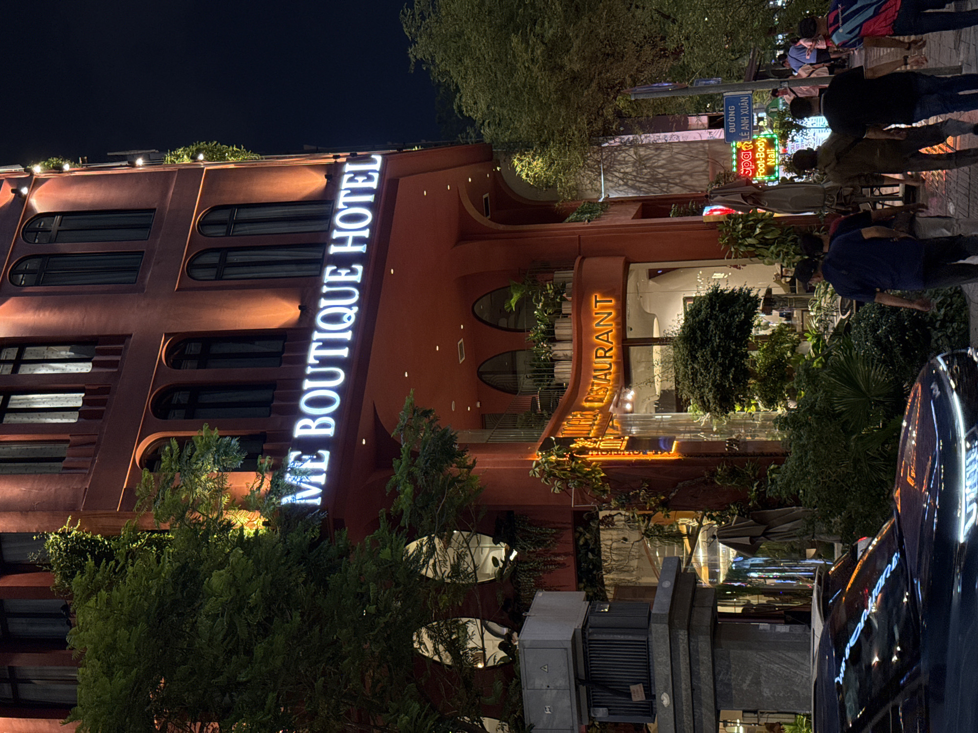 The image shows a nighttime view of a building in Thủ Đức, Vietnam. The building, identified by a prominent sign as "ME BOUTIQUE HOTEL," is of a red-brown hue with arched windows and architectural features. The hotel's restaurant is also lit. People are visible on the sidewalk adjacent to the building, engaged in walking. Street signage, including "Spa Lê Anh Xuân," is present, and a portion of a car is visible. Vegetation, including trees and greenery, is integrated into the hotel's architecture and the surrounding streetscape. Electrical infrastructure is also seen.