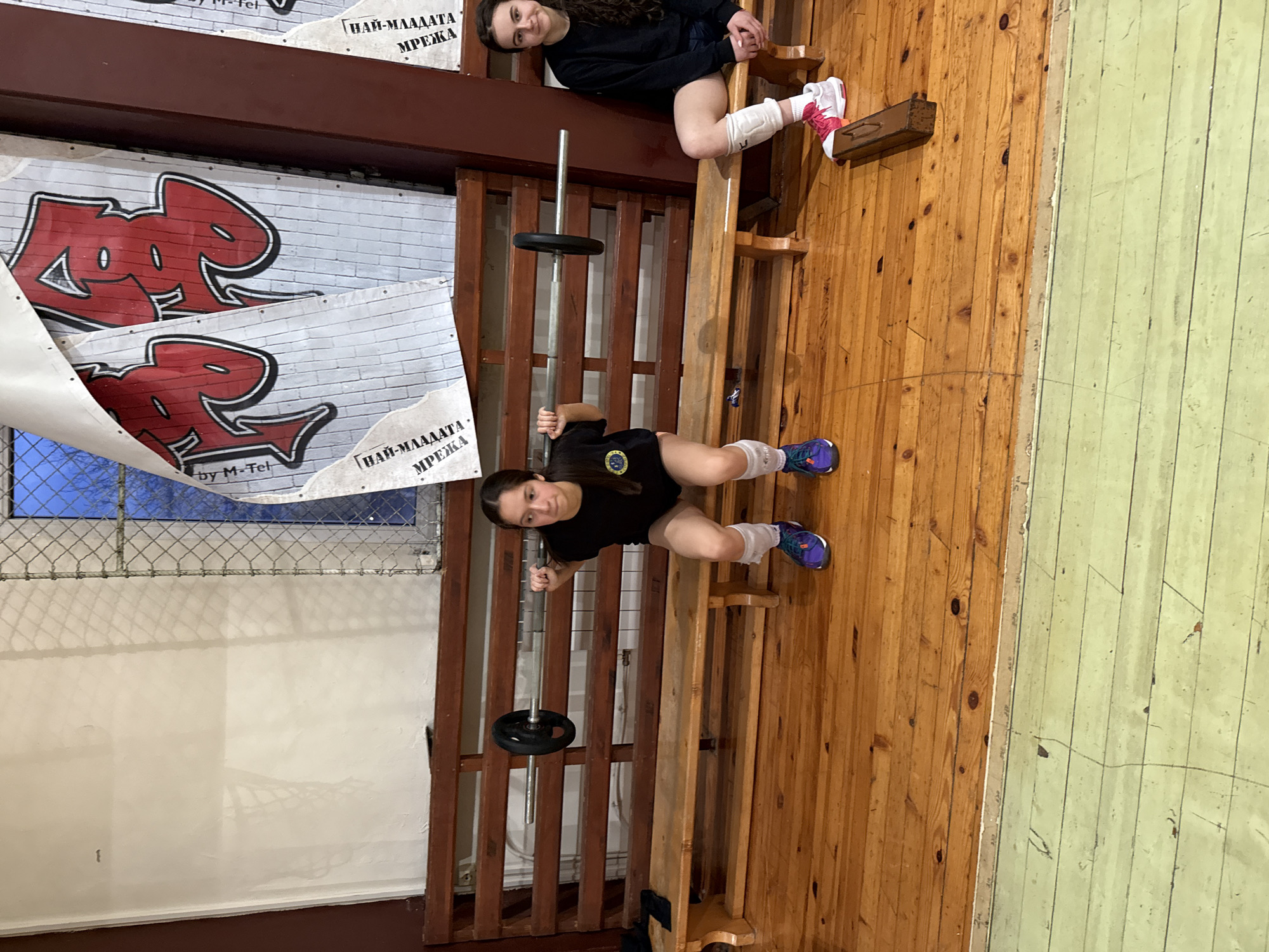 A gymnasium interior in Sofia, Bulgaria, shows two young women positioned atop elevated wooden bleachers. One woman, seated, wears a black top, shorts, and white leg supports, paired with pink and white sneakers. The other woman is prone with a barbell resting above her chest; she is dressed in a black t-shirt, shorts, and white leg supports, completed with blue and purple sneakers. Supporting the barbell is a wooden bleacher structure. Behind the bleachers are two banners reading “Най-младата мрежа by M-Tel” with a red graffiti-style image. A fence partially obscures a window in the background. The right side of the image shows a wooden floor adjacent to a painted section.
