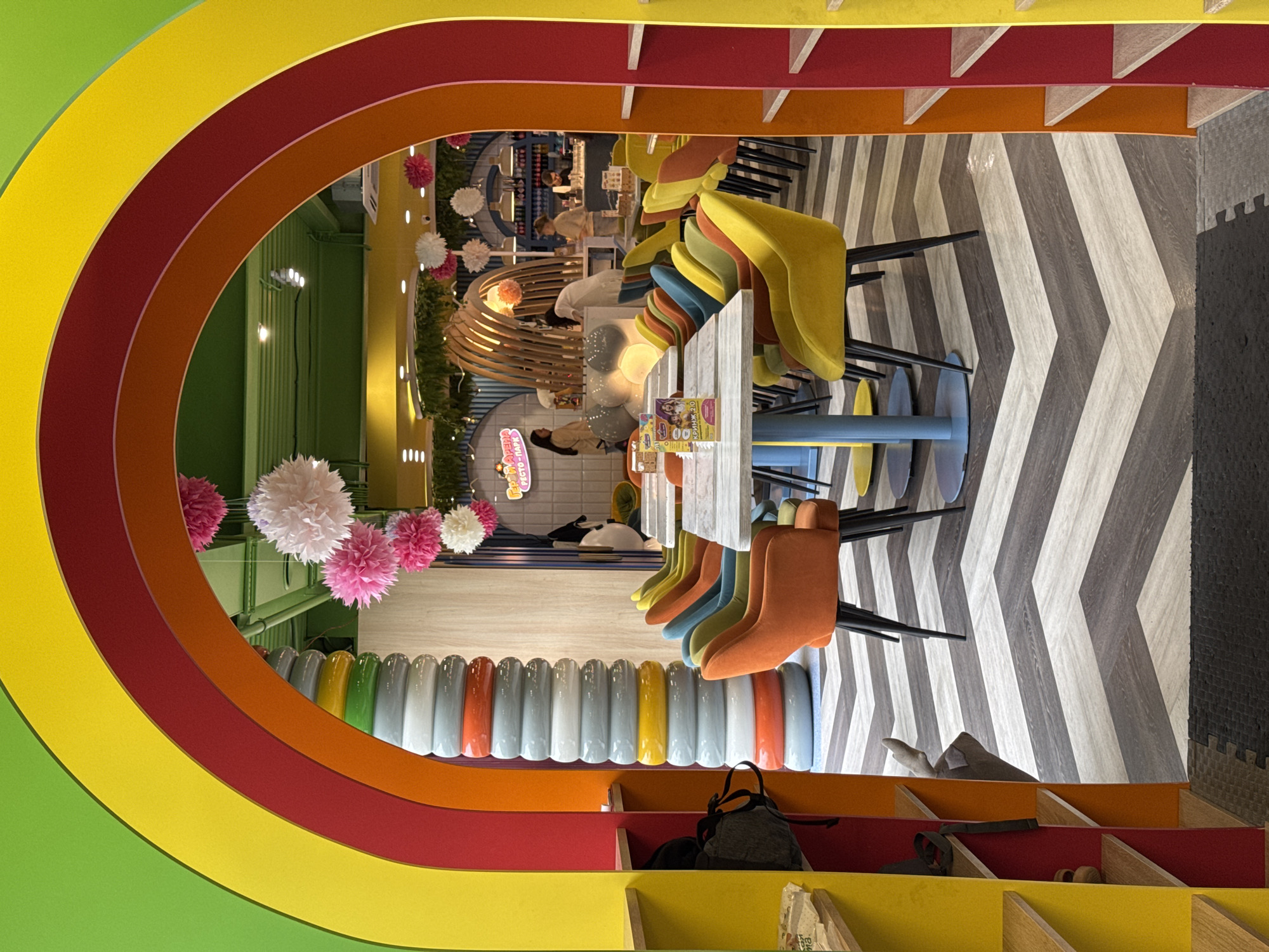 The image depicts an interior space, possibly a children's play area or cafe, located in Sochi, Russia. The scene is framed by a large, colorful arch resembling a rainbow. The arch consists of sections colored green, yellow, red, and orange. The interior space features a chevron pattern floor in shades of gray and white. There are tables with multi-colored chairs, some upholstered in bright yellows, blues, and oranges. A decorative wooden structure, potentially a playscape, is visible in the background. Paper flower decorations hang from the ceiling. Additionally, there is built-in shelving along the bottom of the frame, and some personal items like bags and shoes are visible on the shelves. The general atmosphere is bright and playful, suggesting a family-friendly environment.