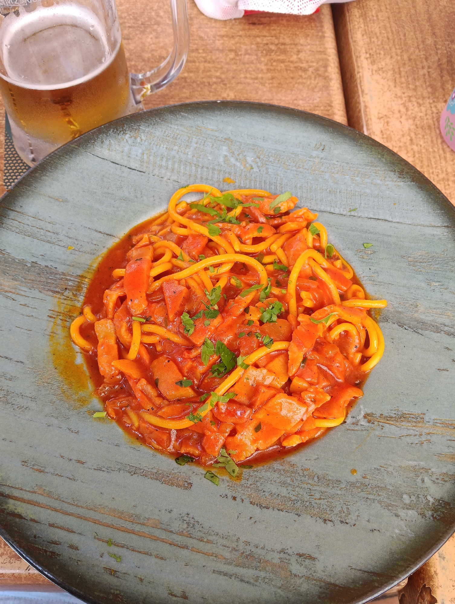 The image captures a close-up view of a meal set on a wooden table, likely at a casual dining establishment in Puerto Del Carmen, Spain. The central focus is a round, rustic blue-grey plate holding a generous serving of pasta. The pasta appears to be a spaghetti-like variety, perhaps slightly thicker or irregularly shaped, with a yellowish-orange hue, thoroughly coated in a vibrant red sauce. This sauce contains chunky pieces of what look like red bell peppers or similar vegetables. Freshly chopped green herbs, possibly parsley or cilantro, are sprinkled over the pasta, adding a touch of garnish.
To the upper left of the plate, a large, clear glass mug of beer is visible. The mug is frosty and covered in condensation, suggesting the beer is very cold and refreshing. The beer itself is a golden amber color with a visible head of foam. The plate and beer mug rest on a light brown wooden table with a distinct grain pattern. A dark-patterned coaster is partially visible under the beer mug. In the background, hints of a white and red patterned fabric (possibly a napkin or tablecloth) and a small, pink object are discernible. The bright, natural lighting indicates it is daytime, and the cold beer suggests a warm climate, fitting for a Spanish holiday destination. The overall scene conveys a relaxed and enjoyable dining experience. No discernible text is visible in the image.