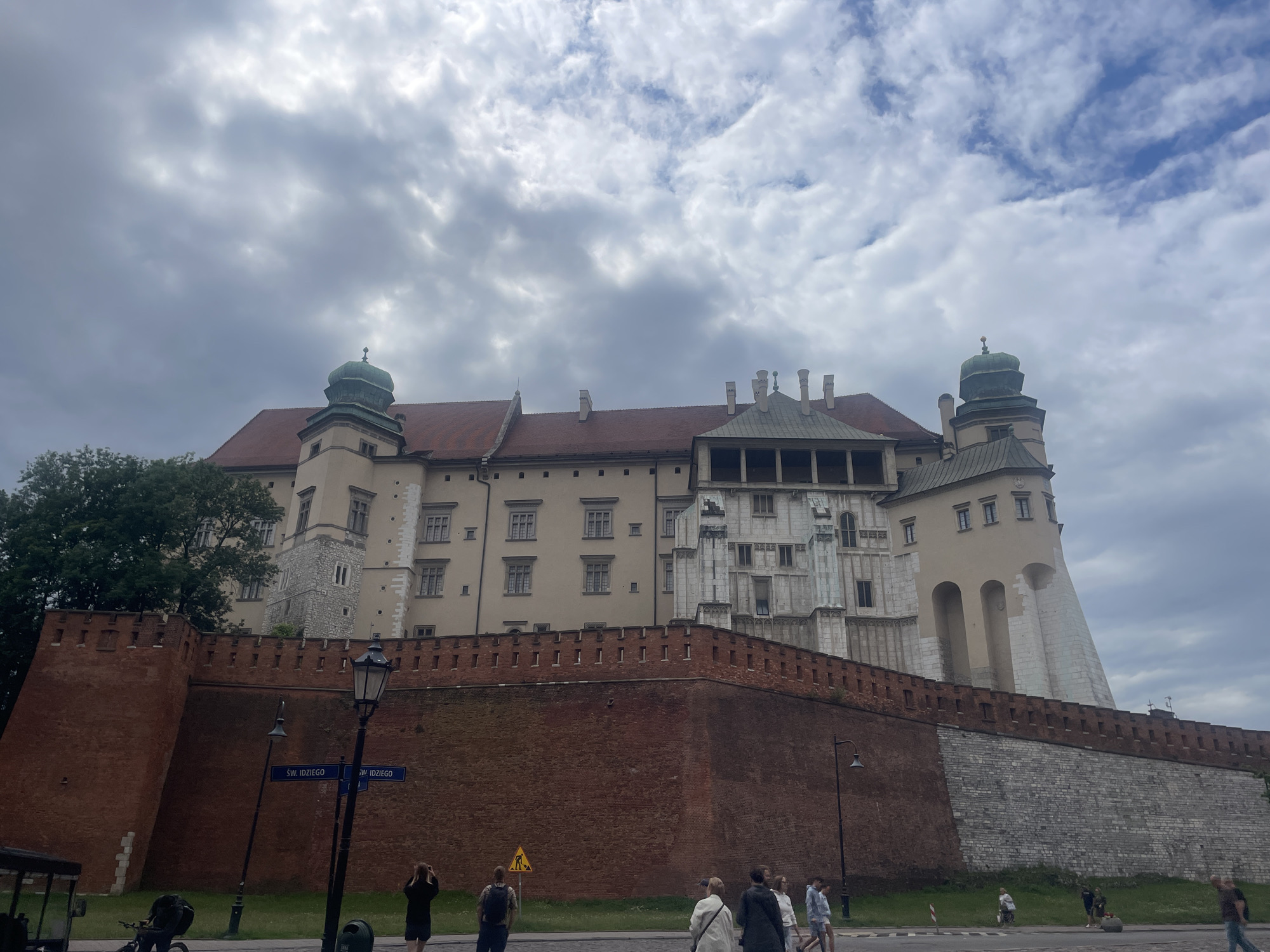 This is a daytime, eye-level, exterior shot of Wawel Castle in Krakow, Poland, under a cloudy sky. The large castle complex features terracotta tiled roofs and light-colored stone walls, with prominent green domed turrets. A tall, red brick wall, topped with battlements, encloses the castle grounds. In the foreground, a street runs along the base of the wall. Several people are visible on the sidewalk and street, some walking and others standing. One person is taking a photograph of the castle. Street signs indicating "ŚW. IDZIEGO" are visible. A lamppost stands near the wall, and a yellow construction sign with a figure is also present. The environment appears to be urban, with the castle as the central subject.