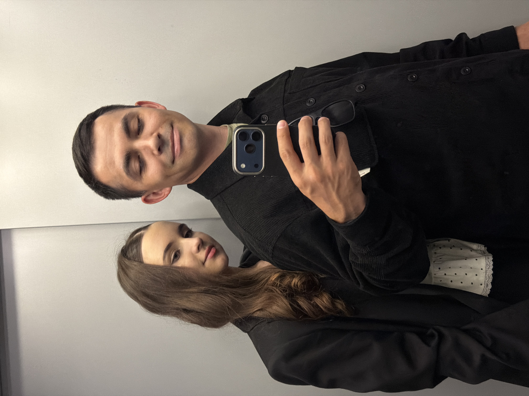 A man and a woman are posing for a selfie in what appears to be a small, enclosed space, possibly a public restroom. The man is in the foreground, holding a smartphone with a visible triple-lens camera in his right hand, and is wearing a dark corduroy-like button-down shirt, with a pair of glasses tucked into the front. The woman is behind him and to his left, wearing a dark blazer over a polka-dotted blouse. The background is a plain, light-colored wall. The location of Izhevsk, Russia cannot be verified from this image.