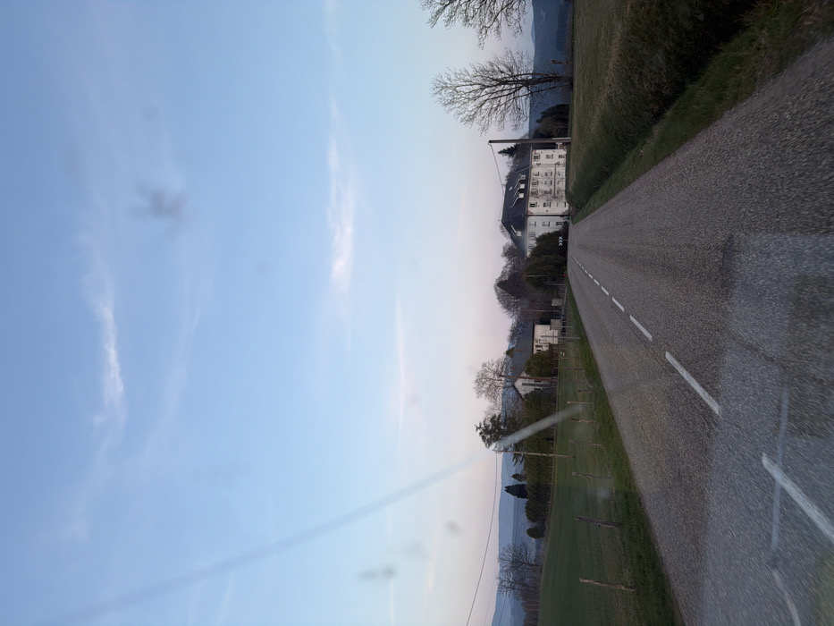 This image depicts a rural road leading into a settlement within Le Saulcy, France, under conditions suggesting twilight. The asphalt road, running from the foreground towards the background, is marked with a continuous white line on its left edge and a dashed white center line.
On the left side of the road, a grassy field is segmented by a fence composed of wooden posts and wire. Beyond this field, a cluster of deciduous trees in a bare state for the season, and at least two structures, one a white building with a dark roof, are visible amidst further foliage. Rolling hills are visible on the distant horizon.
On the right side of the road, a grassy verge separates the roadway from a large, open field. Further down this side, a multi-story white building with a dark, complex pitched roof stands prominently. This building has multiple windows and appears to be a substantial structure. To its immediate right, a road sign with a black arrow on a white background points left, indicating a directional change in the road. Bare deciduous trees are present both in the foreground and surrounding the building. A utility pole stands near the white building.
The sky is predominantly light blue, transitioning to a pale pink-purple hue at the horizon. Wispy white clouds are scattered across the upper sky. A prominent diagonal streak of light glare traverses the image from the upper left to the lower right, and several circular lens flares are also present. No individuals or vehicles are visible in the scene.