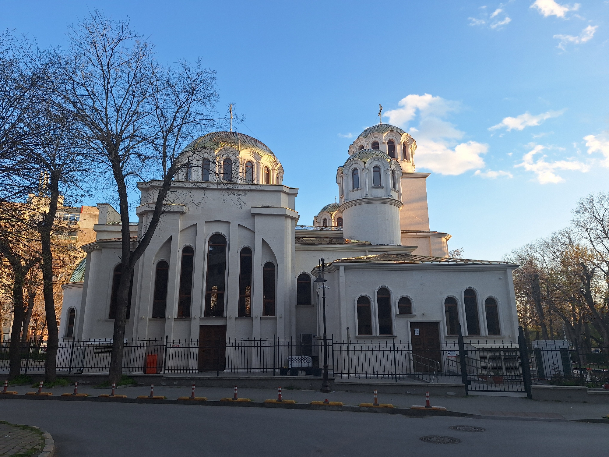An Orthodox church, distinguished by its white facade and multiple golden domes topped with crosses, stands under a clear blue sky with scattered white clouds. The building features various architectural elements including arched windows, a prominent main dome on the left, and a taller, round turret-like structure with its own smaller dome on the right.
In the foreground, several bare deciduous trees, indicative of late winter or early spring, frame the scene, with one large tree dominating the left side. A black metal fence separates the church grounds from a paved street, where yellow and orange traffic bollards are visible, along with manhole covers.
The warm sunlight illuminates the upper sections and domes of the church, suggesting either early morning or late afternoon. No people are visible, and no specific activity is taking place, creating a peaceful, static urban scene. Distant buildings can be glimpsed through the tree branches. A small sign next to a brown door on the church's lower level shows the text "W-M". The setting is an urban environment, in Varna, Bulgaria.