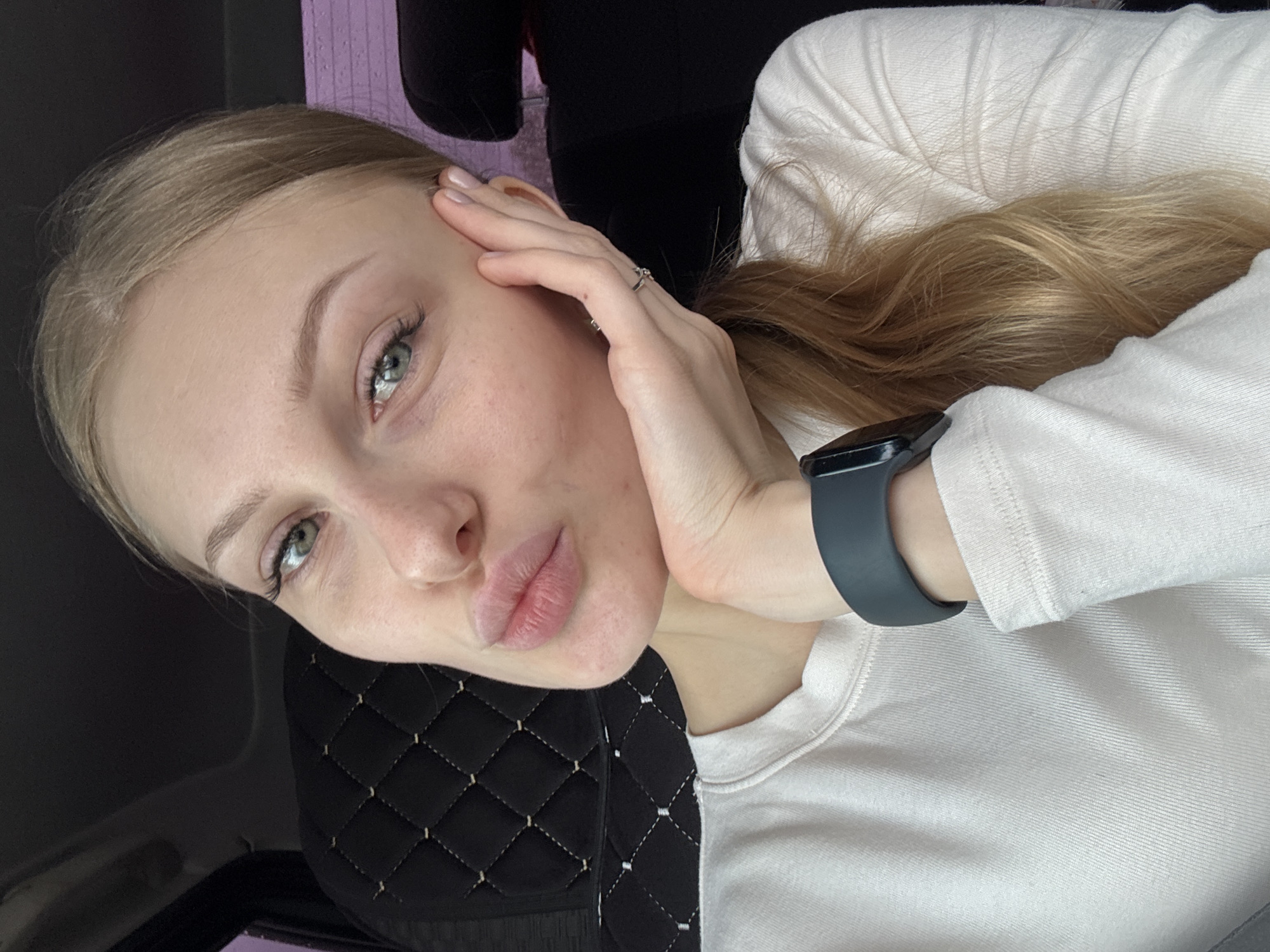A young woman with blonde hair and green eyes is posing for a selfie. She is wearing a white long-sleeved shirt and a dark-colored smartwatch on her left wrist. She is holding her left hand to her face, with a ring visible on her finger. The background consists of a black, diamond-patterned material and a glimpse of a purple surface. The location is Perm, Russia.