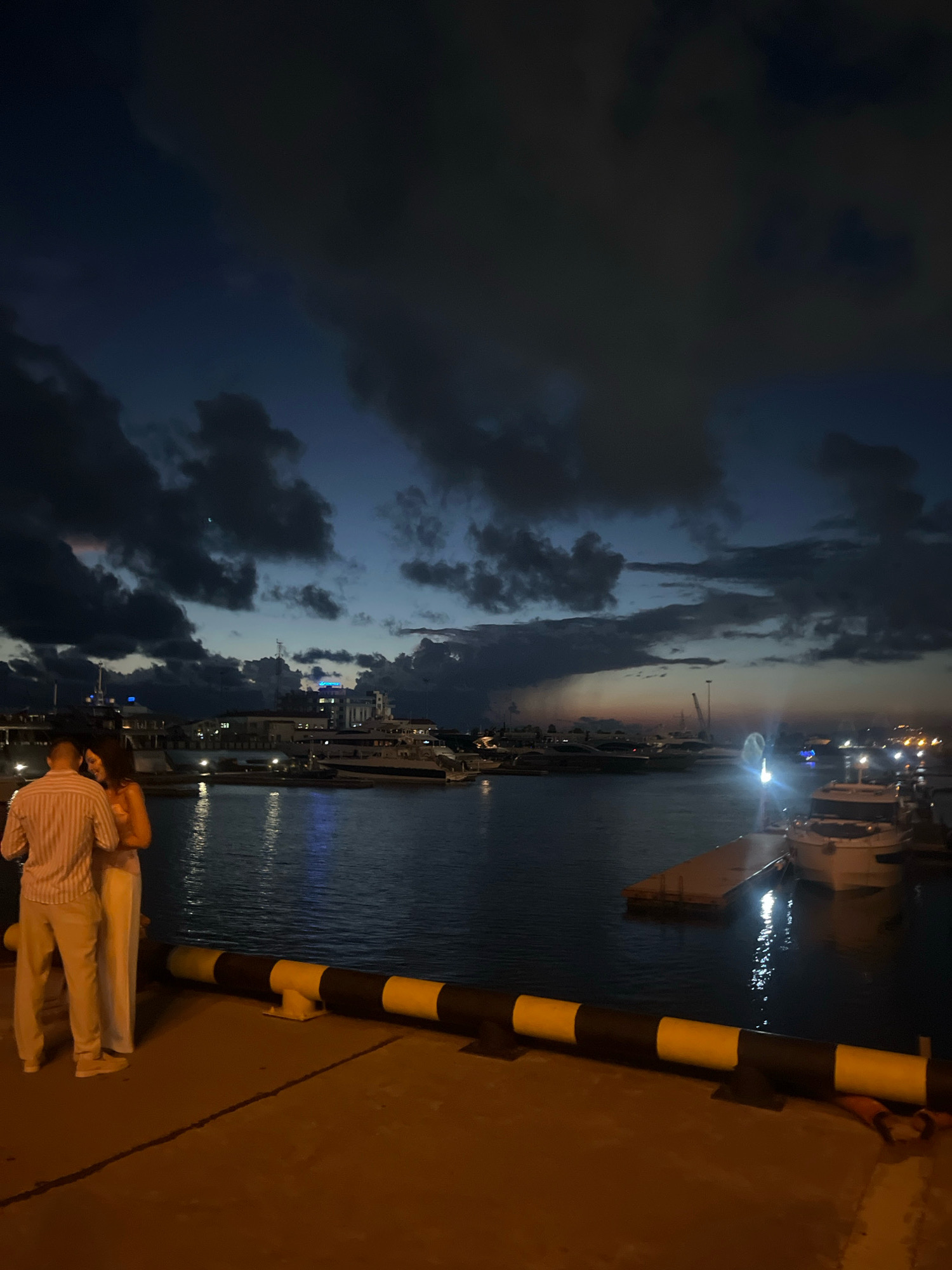 A couple stands close together on a pier at night, facing a harbor filled with yachts and illuminated buildings. The sky above is dark and cloudy, with hints of twilight. The water reflects the lights from the boats and buildings. The couple, one in a striped shirt and light-colored pants, the other in a light-colored dress, appear to be embracing or in conversation. The pier has a striped yellow and black barrier along its edge. The location is identified as the seaport of Sochi, Russia.