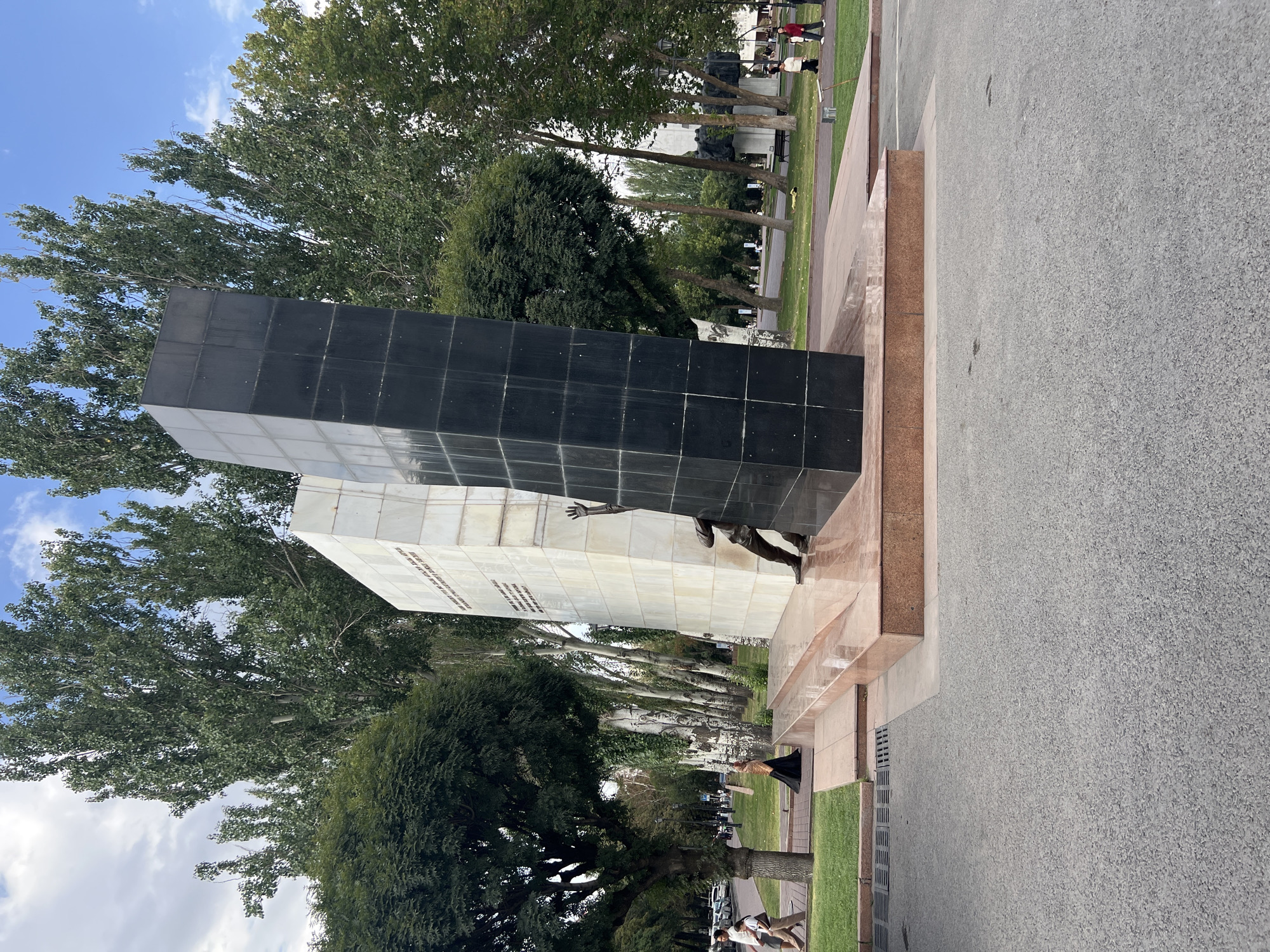 The image depicts a large abstract monument in Bishkek, Kyrgyzstan. The monument is composed of two upright rectangular slabs, one white and inscribed with text, the other black and textured. A bronze statue of a person reaching upwards is attached to the black slab, appearing to climb or emerge from it. The monument is situated on a stepped, rose-colored stone base, surrounded by a paved area and grass. Several trees with lush green foliage line the background, and a cloudy blue sky is visible above. In the middle ground, a few individuals are walking on a paved path near the monument. The foreground shows a grey asphalt surface with some faint tire marks. The overall scene is outdoors, suggesting a public space within the city.
