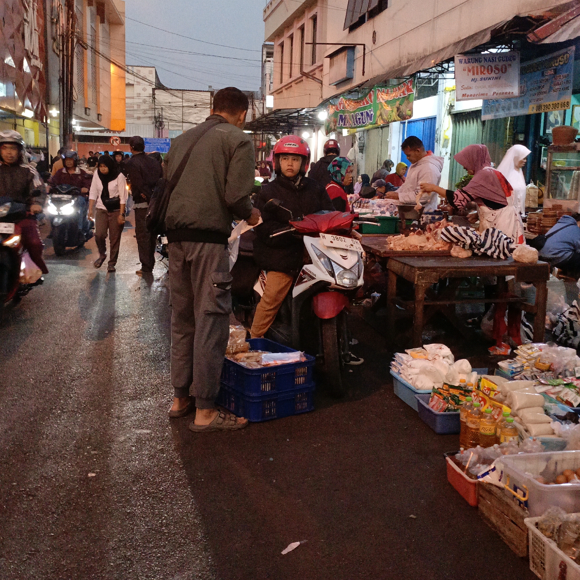 The image depicts a street scene at dusk in Salatiga, Indonesia. The street is wet, suggesting recent rain, with reflections of lights visible. The scene is bustling with activity, likely a market or street food area.
In the foreground, two men are central to the image. One man, facing away from the camera and towards a motorcycle, is wearing a dark green jacket and grey trousers. He appears to be interacting with the other man, who is seated on a motorcycle and wearing a red helmet. The seated man is wearing a dark jacket and brown pants. A white and red Honda motorcycle is positioned between them.
To the left of the main figures, several people are seen walking or on motorcycles. Some are wearing helmets and casual clothing. Further down the street, buildings line the sides, with some illuminated by lights. Signs with Indonesian text are visible on the buildings, one of which reads "WARUNG NASI GUDER 'MIROSO'".
To the right of the central figures, a food stall is set up with various goods displayed. Tables are laden with raw meat, packaged food items, bottles of liquid, and baskets of eggs. Several individuals, including women wearing headscarves, are attending to the stall, possibly serving customers or preparing food. The overall impression is a vibrant, everyday scene of commerce and community life in an Indonesian city.
