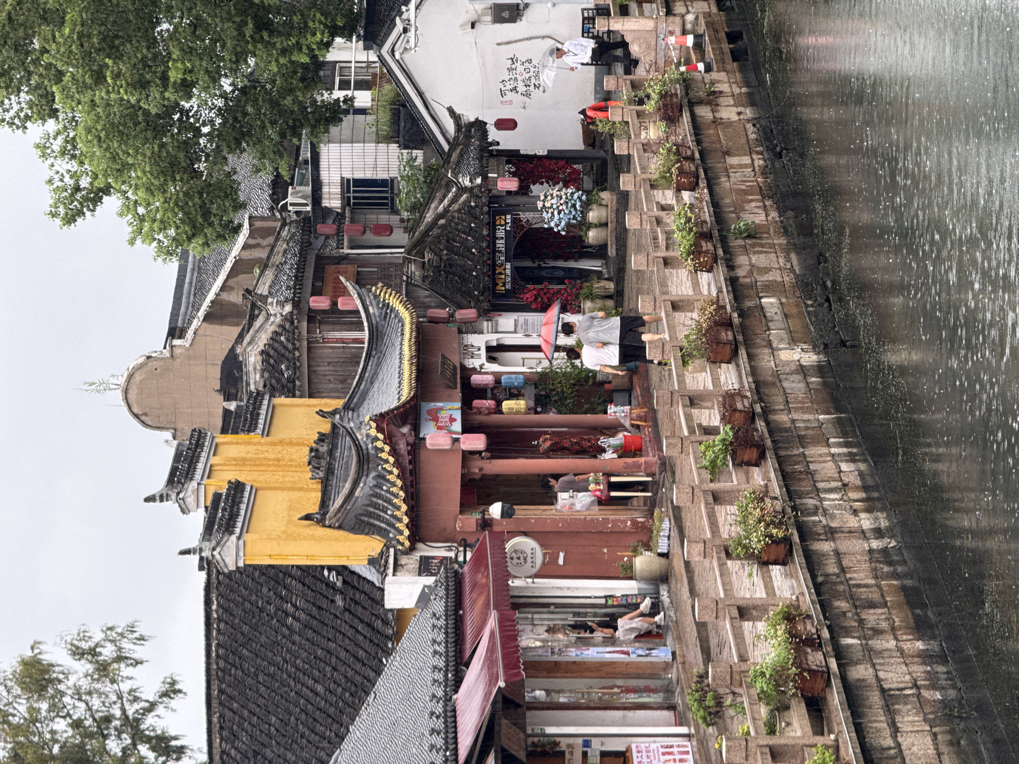 This image depicts a street scene in the city of Qingpu District, China, likely a canal-side promenade lined with traditional Chinese architecture. The buildings feature dark tiled roofs with upturned eaves, many adorned with red lanterns. The architecture is a mix of historic styles, with some facades painted in vibrant yellow and others in white. The street itself is paved and runs alongside a waterway, with a stone embankment and a wooden railing separating the walkway from the water. Planters with greenery are placed along the railing, and several potted plants are visible in front of the shops.
Several people are present in the scene. On the left, a person is sitting on a stool in the doorway of a shop. Further down the street, two individuals are walking together, one holding a red umbrella. To the right, a person in a white shirt is holding a clear umbrella and standing near a wall with Chinese calligraphy. Another person is visible behind a counter inside one of the shops. Various objects are scattered around, including shopping bags, umbrellas, potted flowers, and signage. The overall atmosphere suggests a bustling, perhaps slightly damp, day with people engaged in shopping and strolling along the scenic waterway. The presence of umbrellas indicates it might be raining or recently rained.