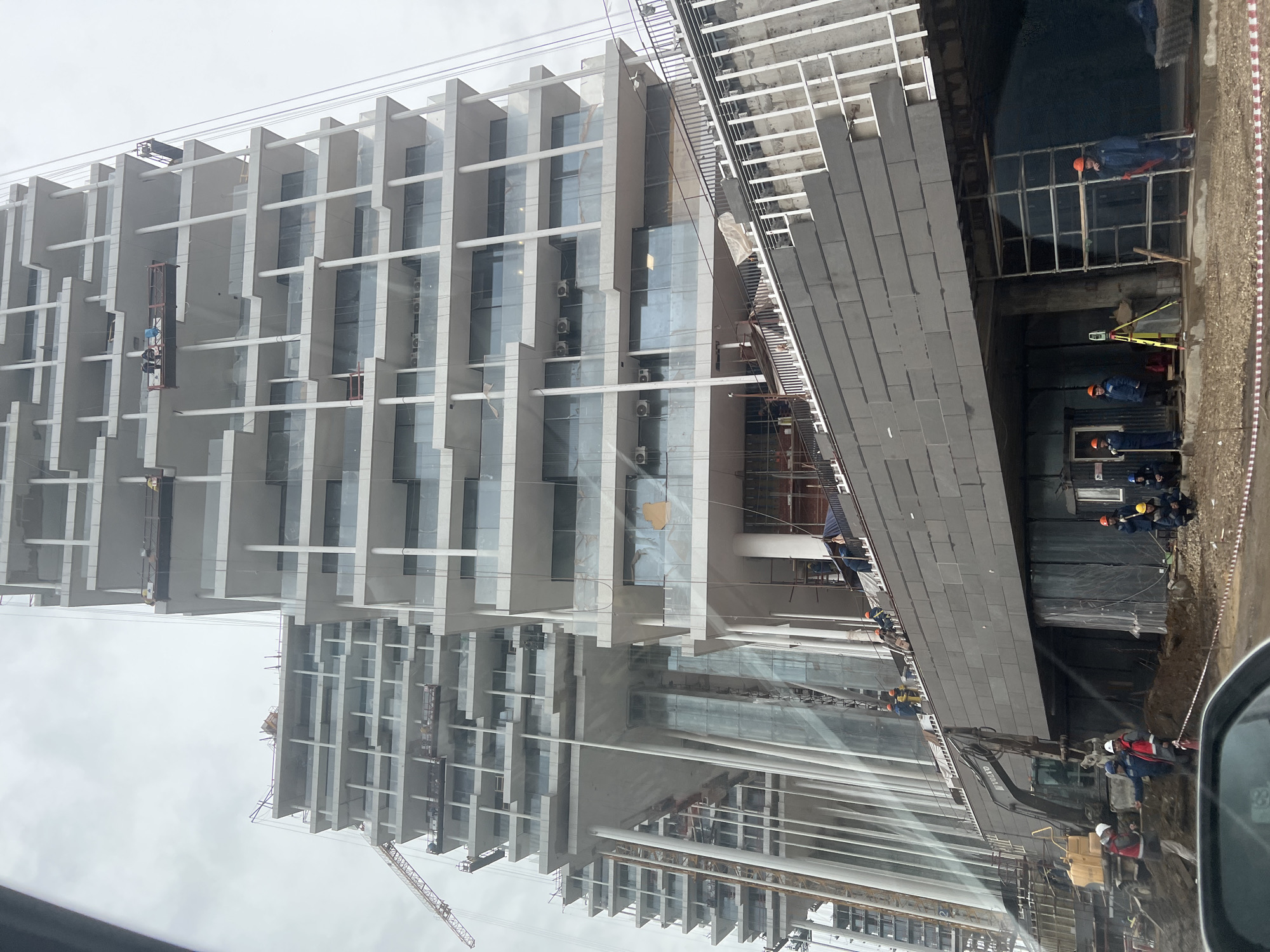 The image shows a construction site in Khimki, Russia. A tall, modern building under construction is visible on the left. The building has a white facade with large glass windows and vertical white beams. Some workers are on a suspended platform performing tasks on the exterior. To the right of the building, a construction crew works on a ground-level project. They are wearing hard hats and working around scaffolding and an excavation site. A construction crane is visible in the background on the left. A car mirror is visible in the bottom right corner, indicating the photo was taken from inside a vehicle. The sky is overcast.
