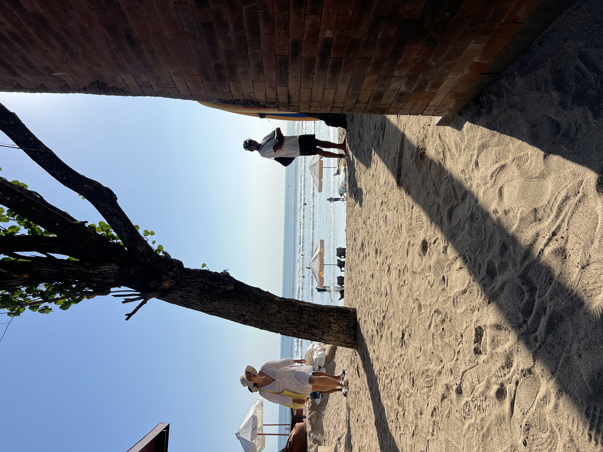 The image depicts a beach scene in Kuta, Indonesia. The foreground is dominated by sand with numerous footprints and the shadow of a tree. In the midground, a woman in a white dress and a hat walks from left to right, carrying a yellow bag. Behind her, a man in a white t-shirt and dark shorts stands with his arms crossed, facing the ocean. Several beach umbrellas, chairs, and surfboards are visible near the water's edge. The ocean is calm with small waves, and a few people are in the water. A brick structure is on the right side of the frame, casting a large shadow. The sky is clear and blue.