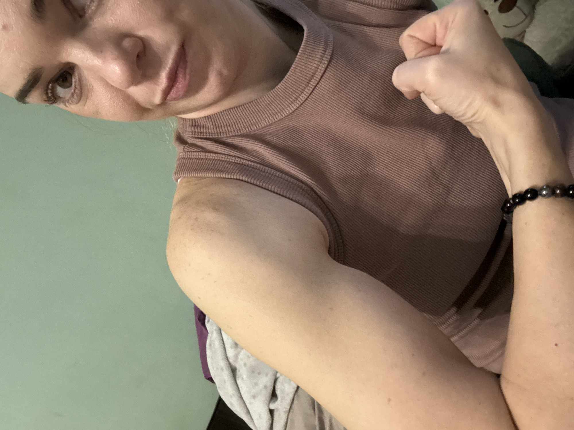 An indoor, waist-up shot shows a woman in a mauve ribbed tank top flexing her arm. She has fair skin with freckles on her arm and shoulder. Her dark hair is pulled back, and she is looking to the right of the camera with a slight smirk. She is wearing a dark beaded bracelet on her left wrist. The background is a solid light green wall. The lighting is dim. The image was captured in Unknown, Russia.