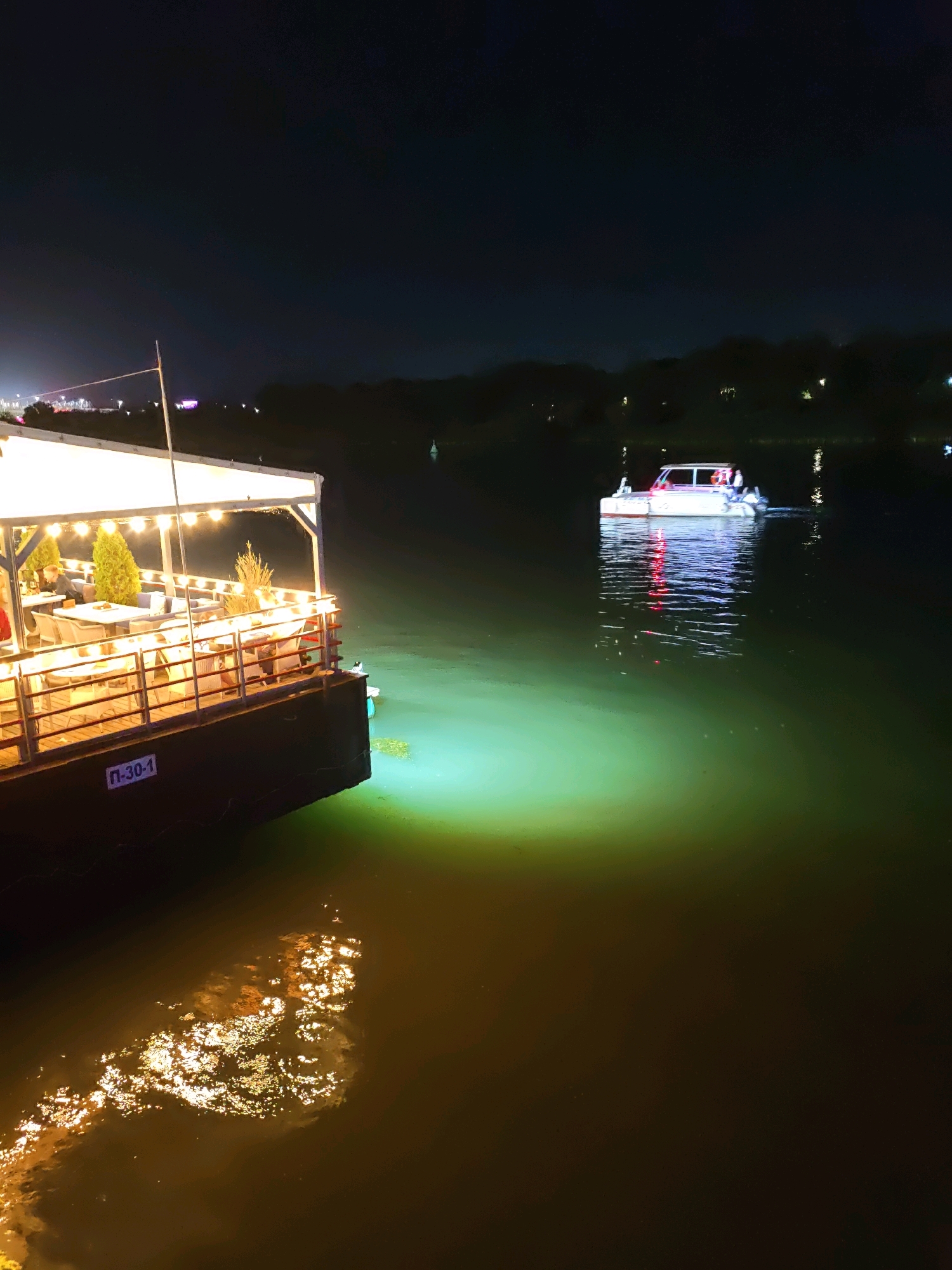 The image depicts a night scene on a body of water, likely the Don River in Rostov-on-Don, Russia. In the foreground, a floating restaurant or cafe is docked, illuminated by strings of warm-toned lights. Tables and chairs are visible on its deck, some occupied by patrons. A portion of the restaurant's hull has a visible designation "П-30-1". The water around the restaurant is lit by a green underwater light, casting a glow. In the middle ground, a white motorboat with red accents is moving away from the viewer, its lights reflecting on the water. The background shows a dark, tree-lined shore with distant city lights. The sky is dark and clear, suggesting nighttime.